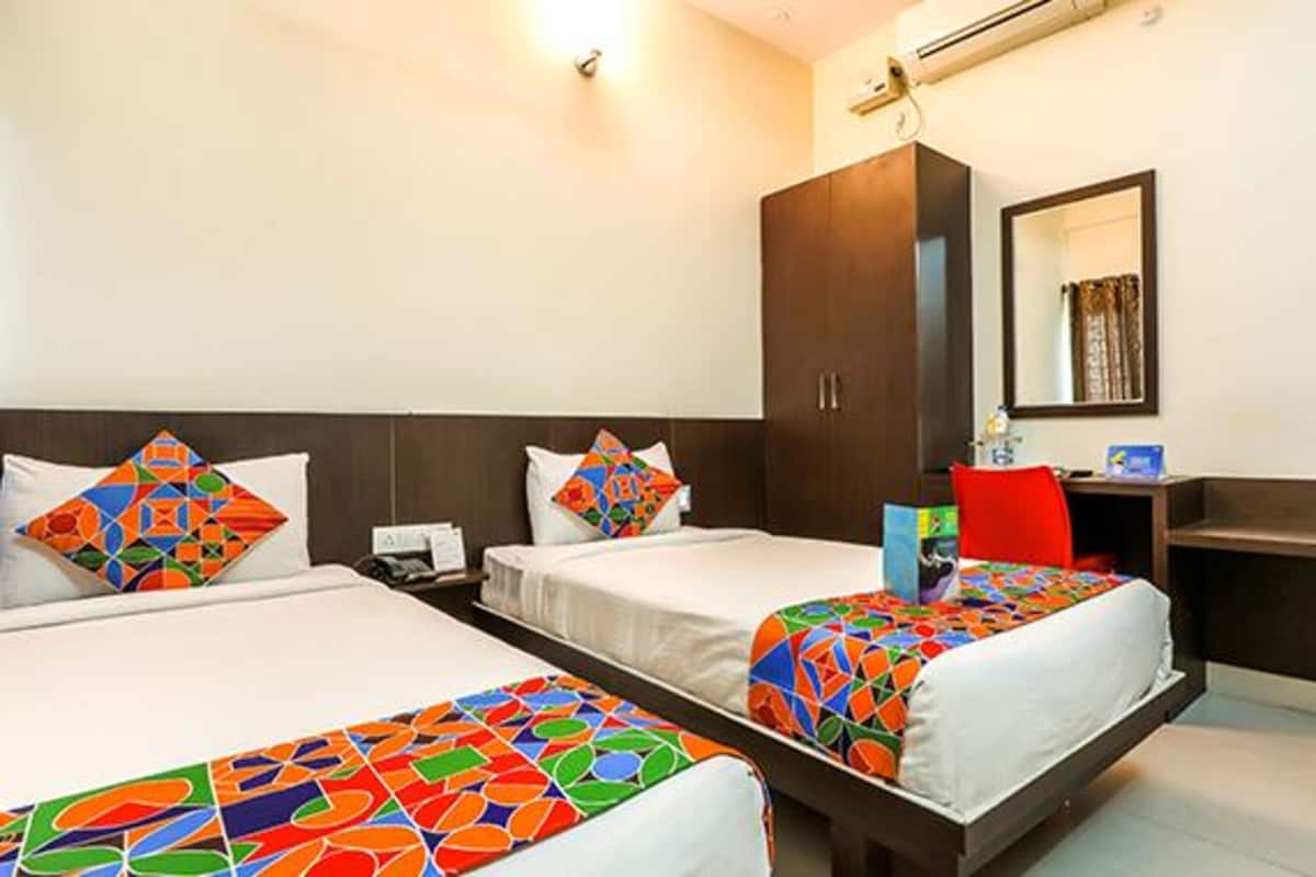 A hotel room with two beds, each with colorful geometric-patterned bedspreads. There is a desk with a chair, a mirror, and a small table with a lamp.