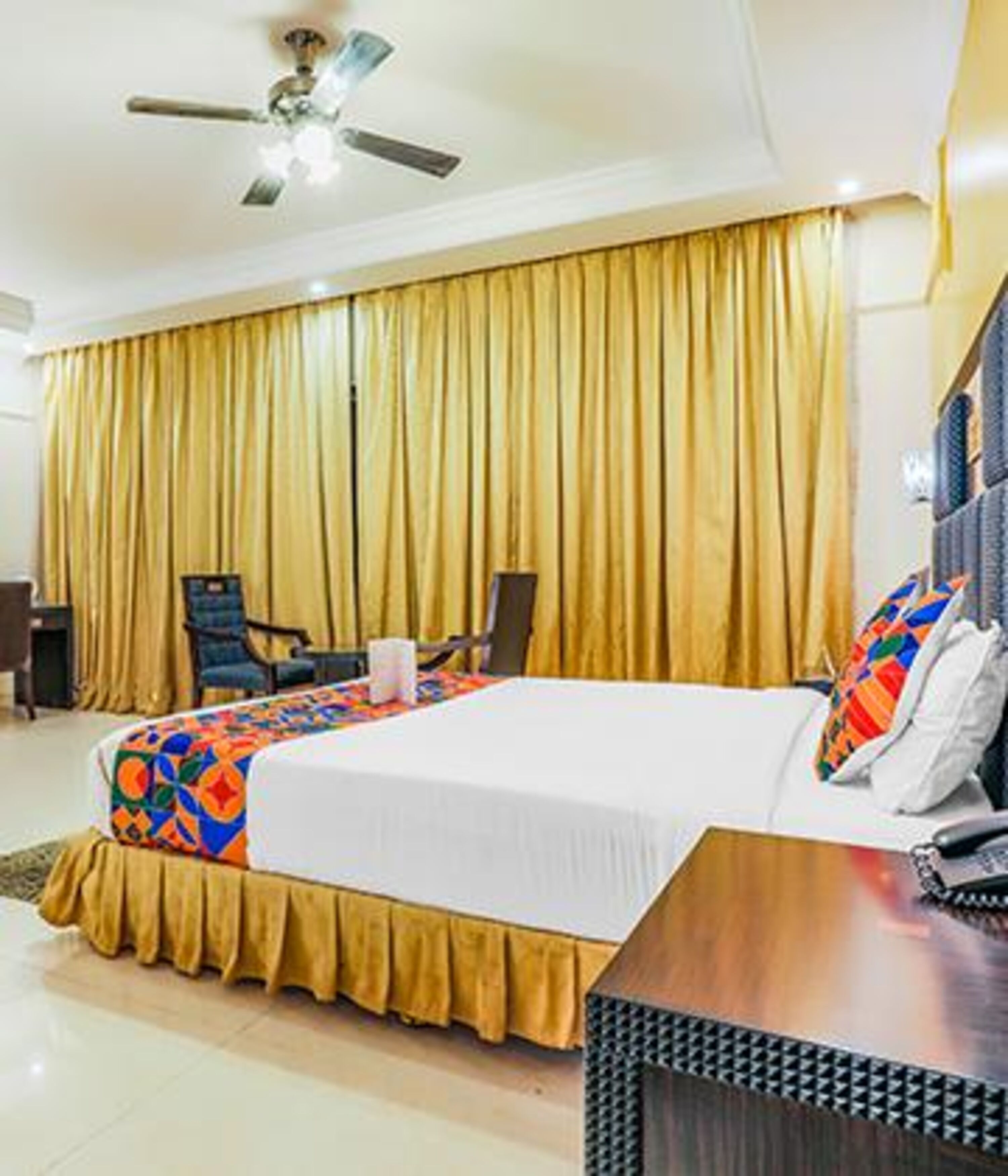 A hotel room with a bed, two chairs, a desk, a telephone, and a ceiling fan.