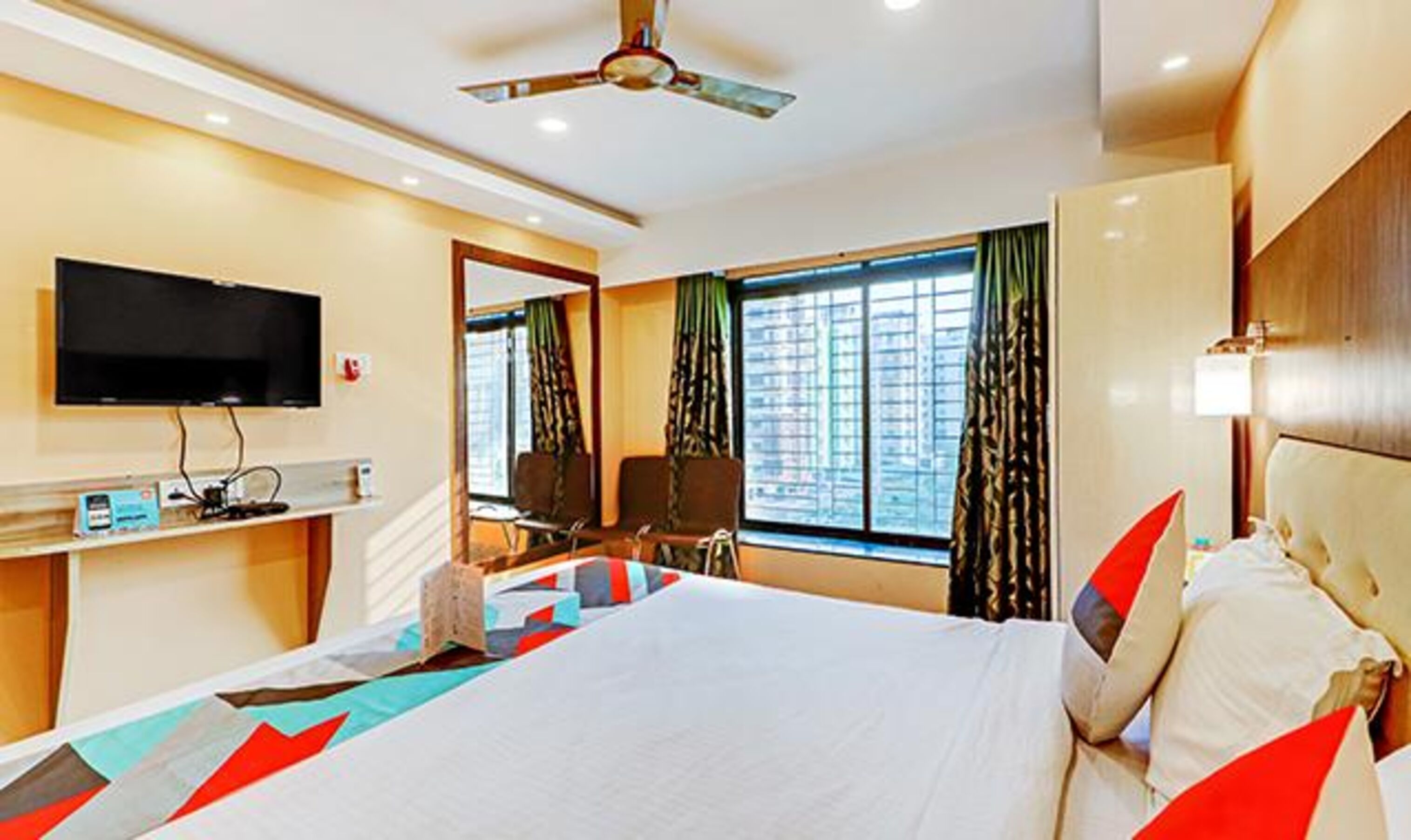 A hotel room with a bed, a television, a desk with a computer, a window with curtains, and a ceiling fan.