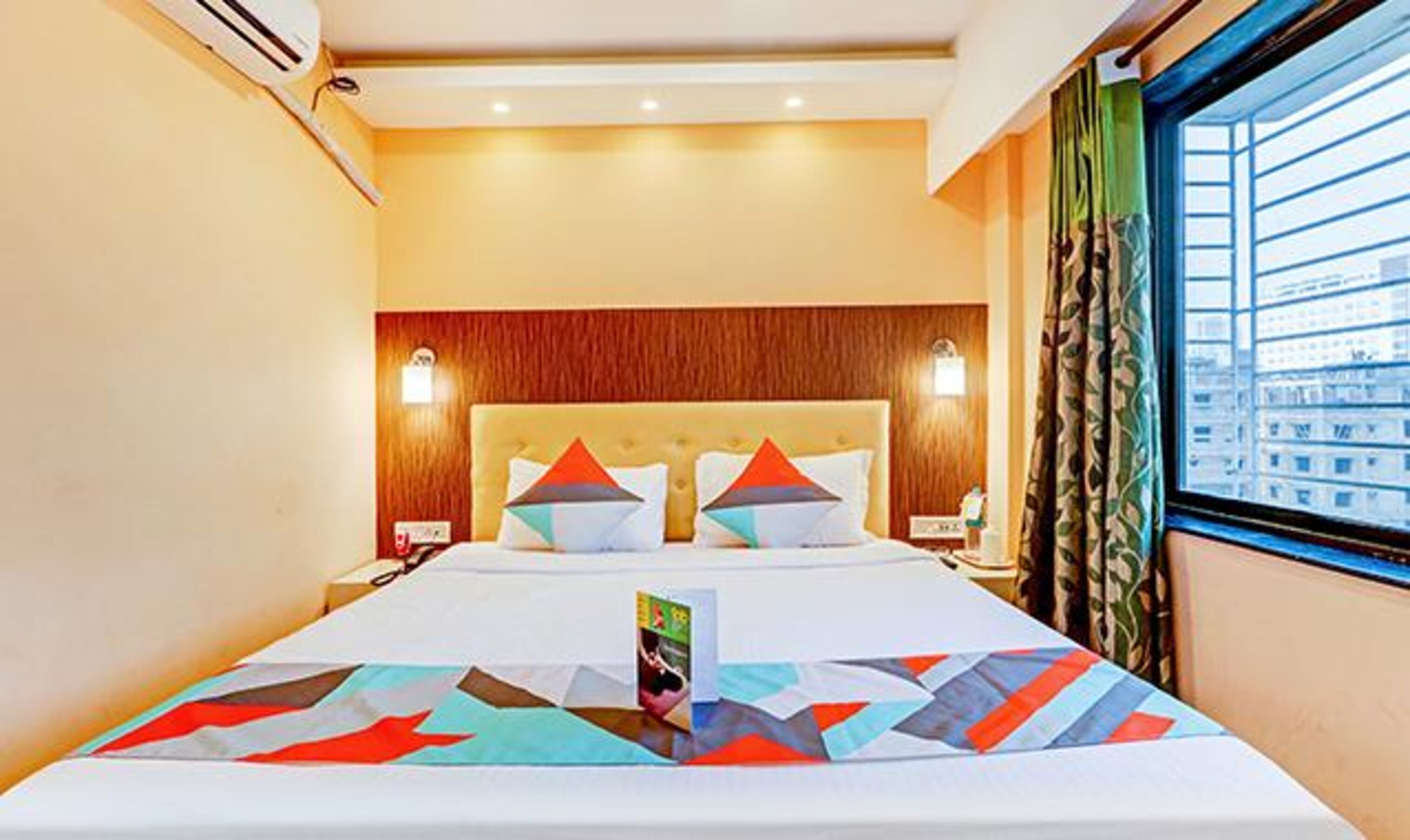 A hotel room with a large bed, a wooden headboard, two bedside lamps, and a window with a view of buildings.