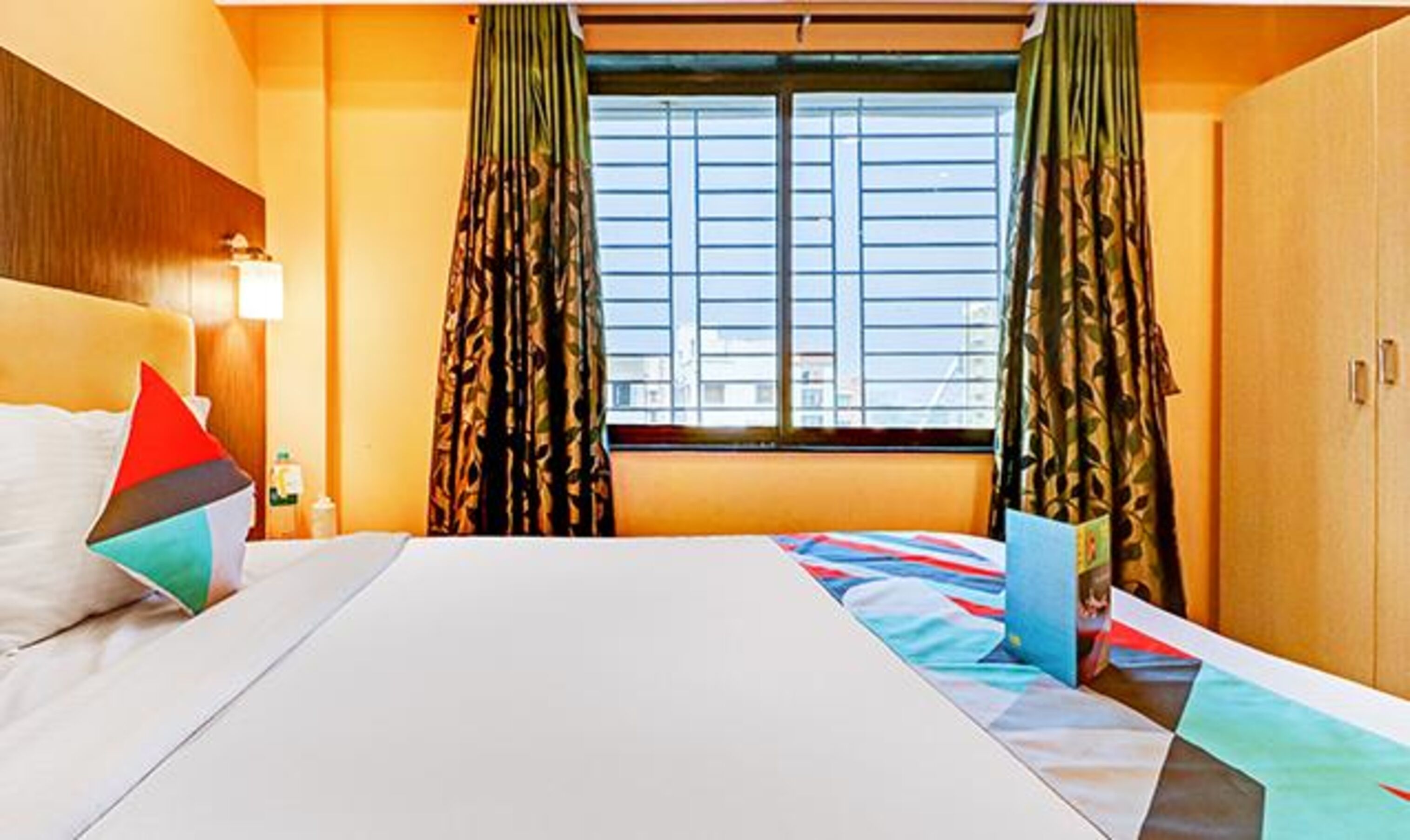 A hotel room with a large bed, two pillows, a nightstand, a window with curtains, and a view of the city.