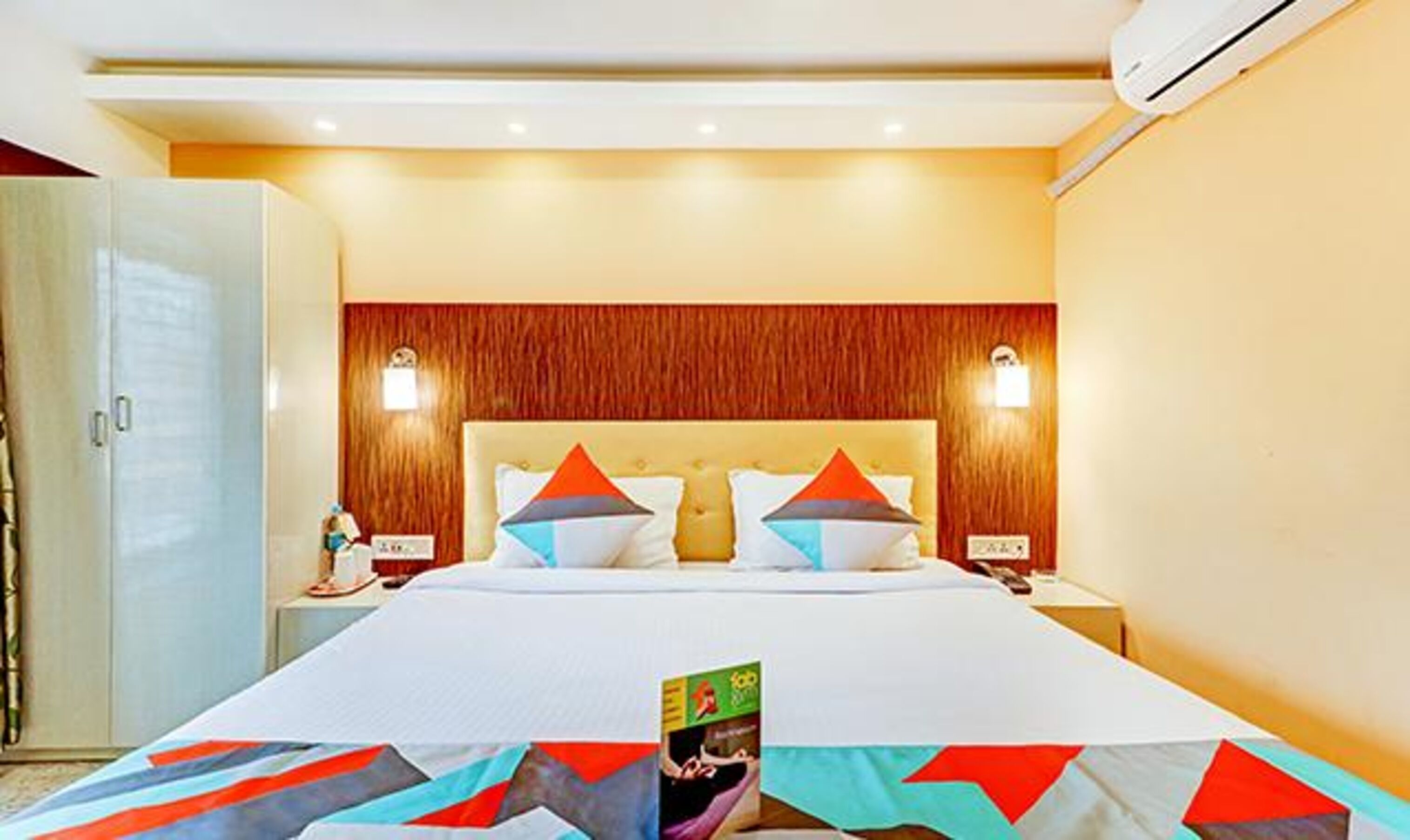 Una habitación de hotel con una cama grande, cabecera de madera, dos mesitas de noche y un equipo de aire acondicionado.