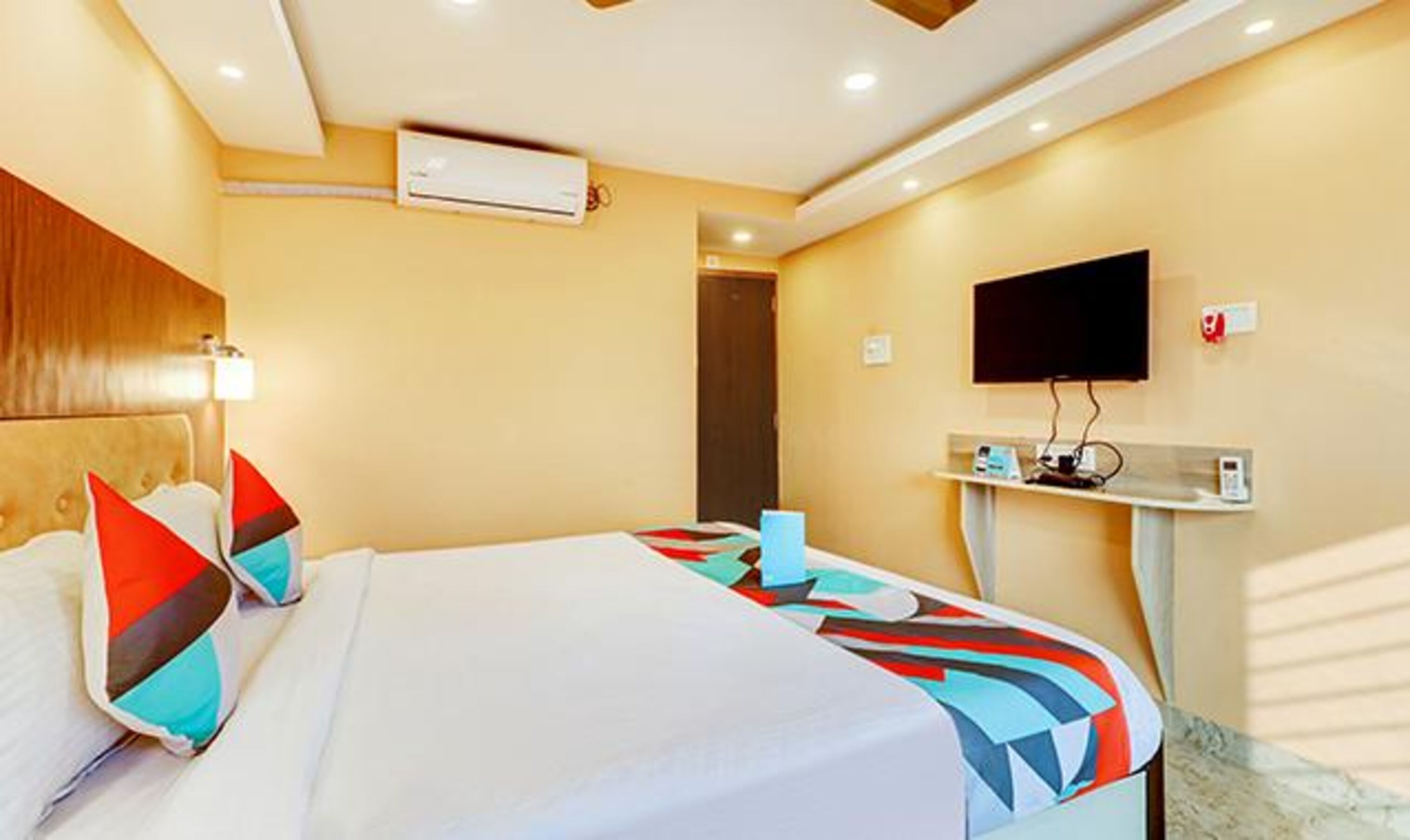 A hotel room with a bed, pillows, a flat-screen TV, a small table, and a wall-mounted air conditioner.