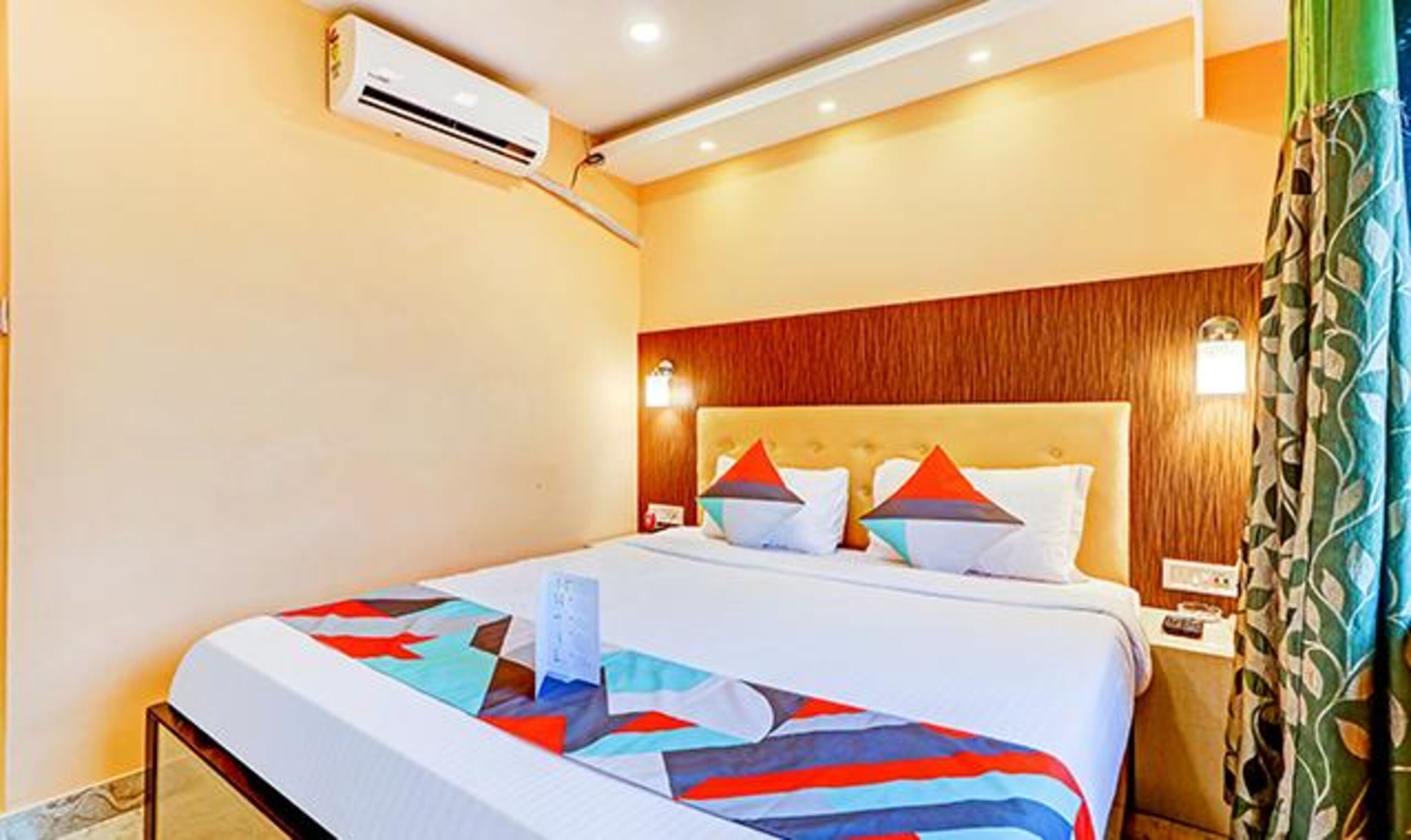 A hotel room with a bed, an air conditioning unit, a nightstand, and a wall-mounted lamp.