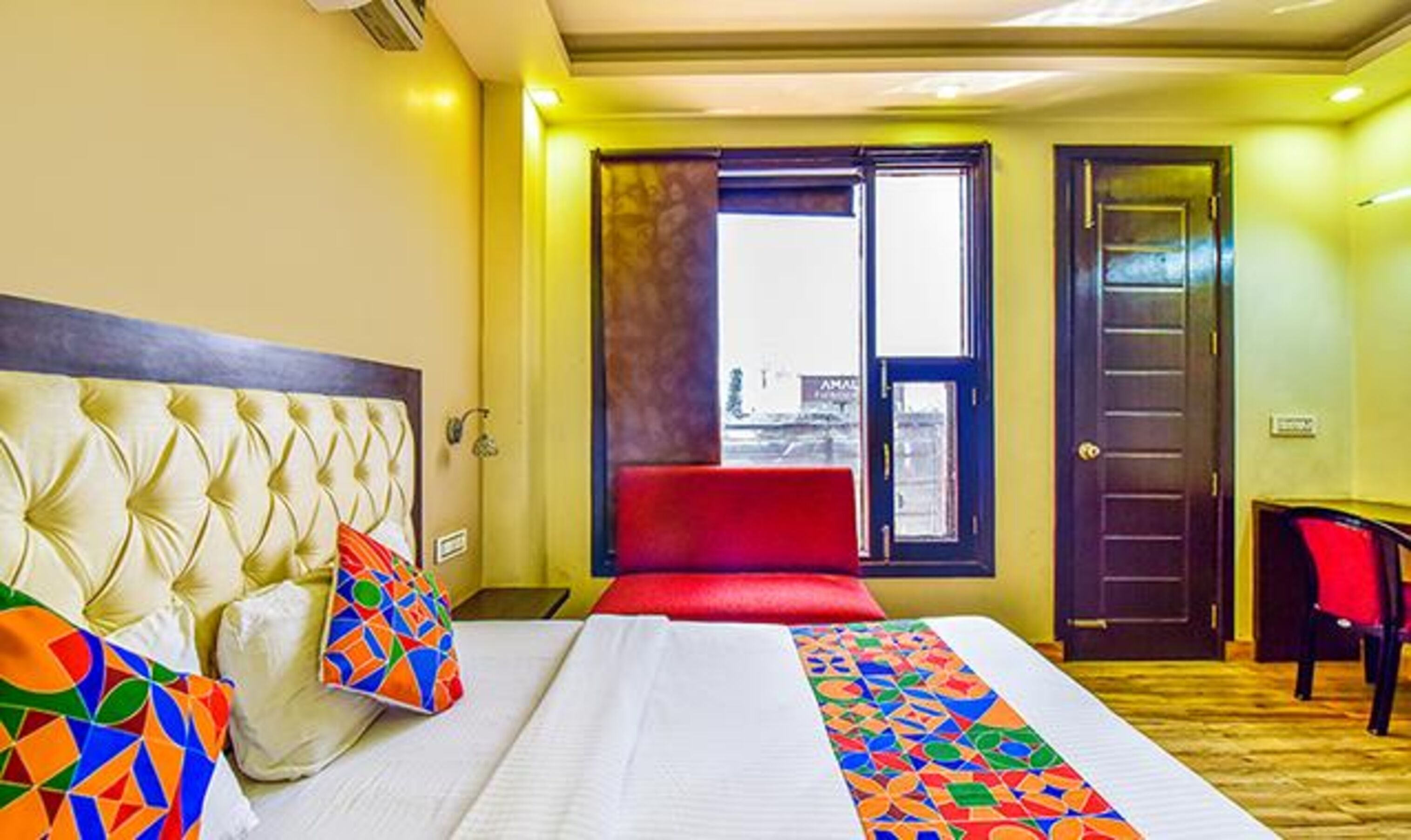 A hotel room with a large bed, a red chair, a desk, and a window with a view of buildings.