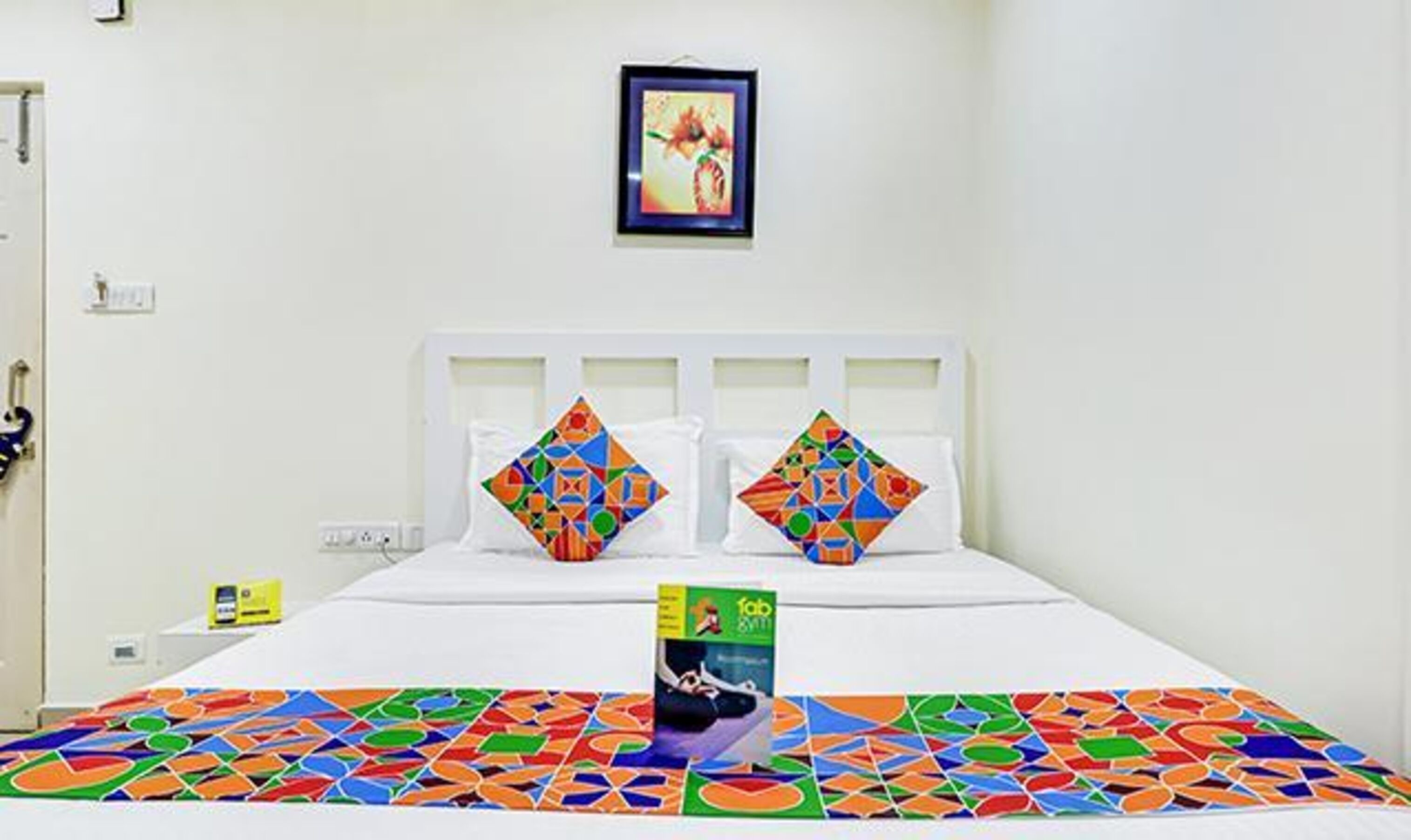 A hotel room with a bed, two pillows with geometric patterns, a framed picture on the wall, and a colorful rug.