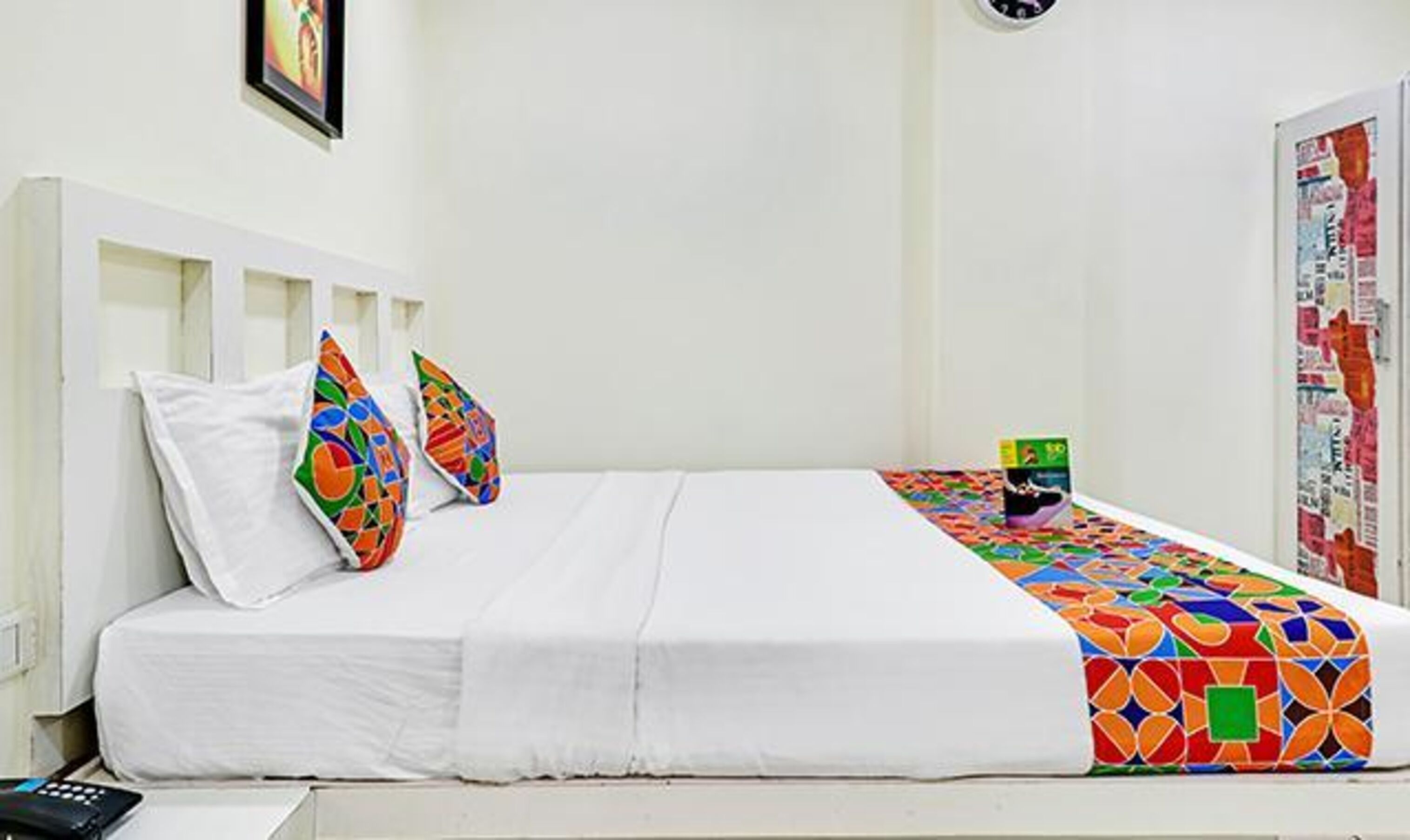 A bed with white bedding and colorful pillows, a small bedside table with a book, and a wall with a poster.
