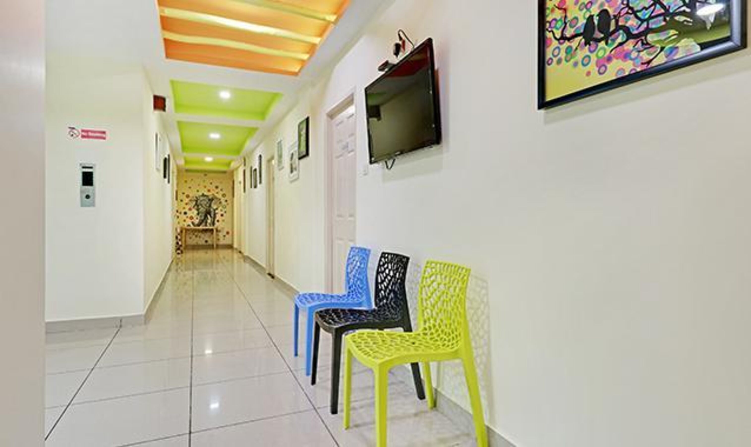 Un pasillo con piso de baldosas, techo verde y televisor montado en la pared. Hay tres sillas en colores azul, negro y amarillo.