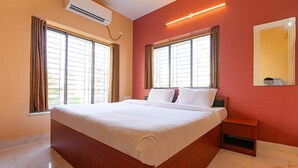 Une chambre d’hôtel avec un grand lit, deux fenêtres avec des rideaux, un climatiseur, une table de chevet et un miroir au mur.