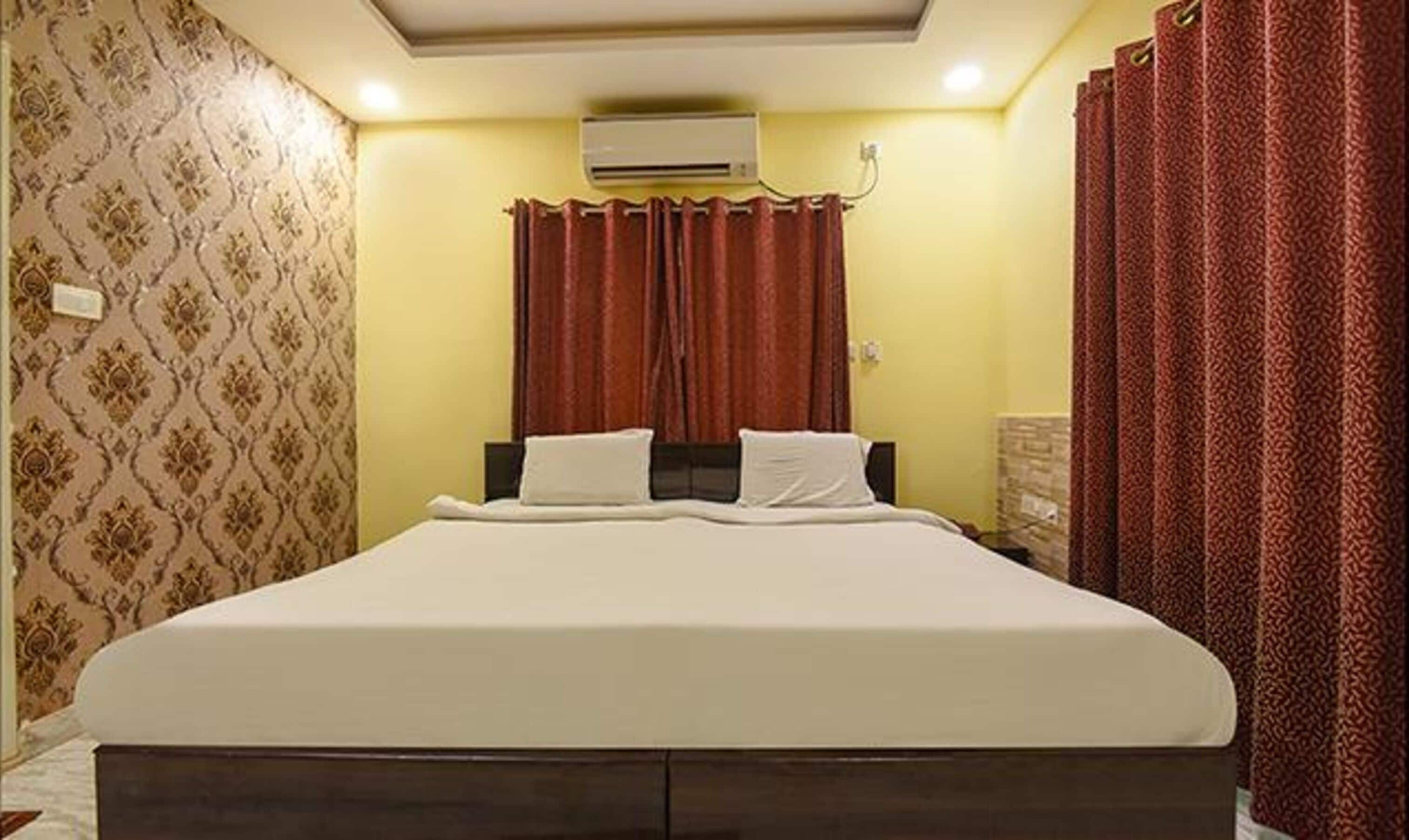 A hotel room with a bed, curtains, an air conditioning unit, and patterned wallpaper.