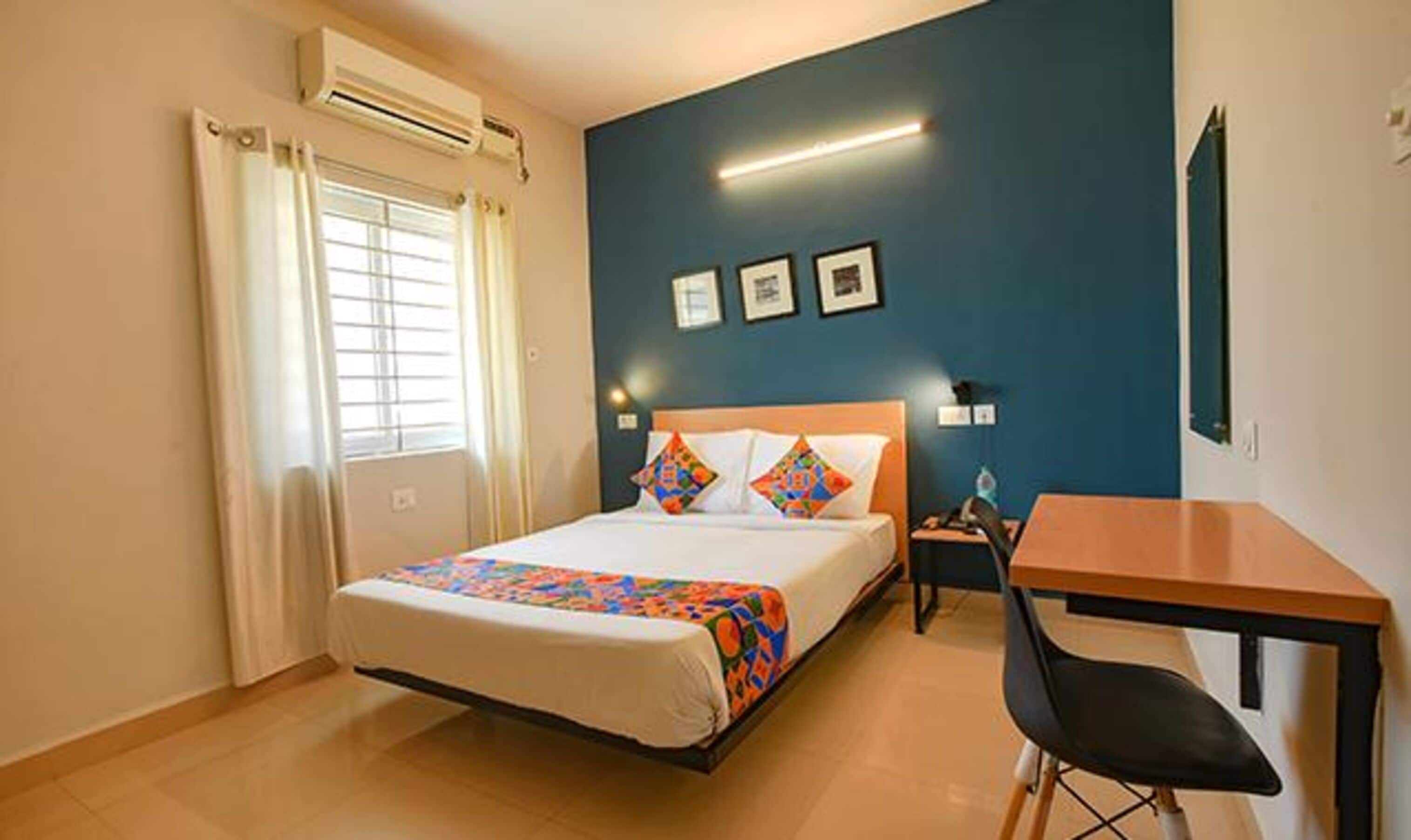 A hotel room with a bed, a desk, a chair, a window with curtains, an air conditioner, and framed pictures on the wall.