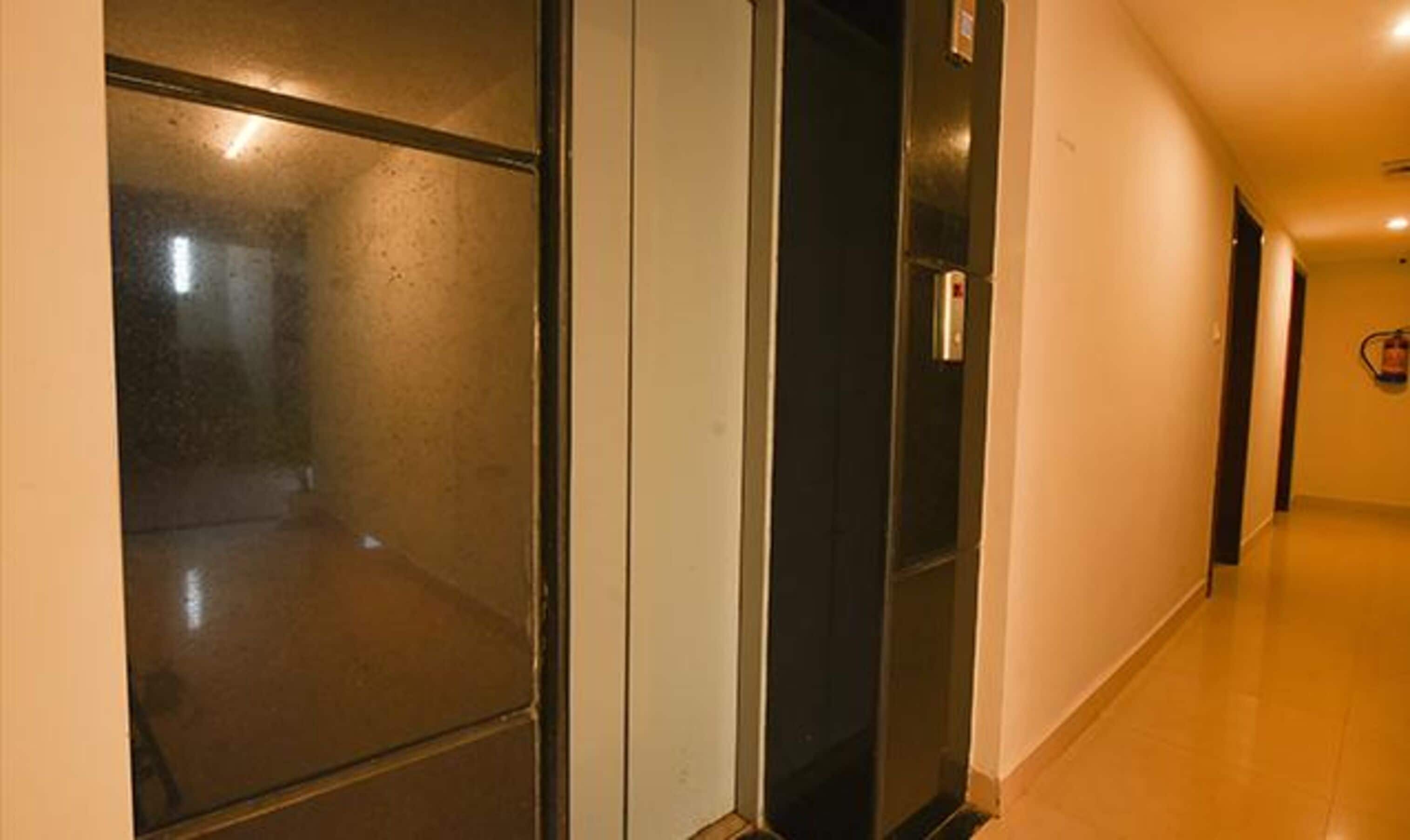 Un pasillo con una puerta de ascensor de vidrio, un detector de incendios y suelo de baldosas.