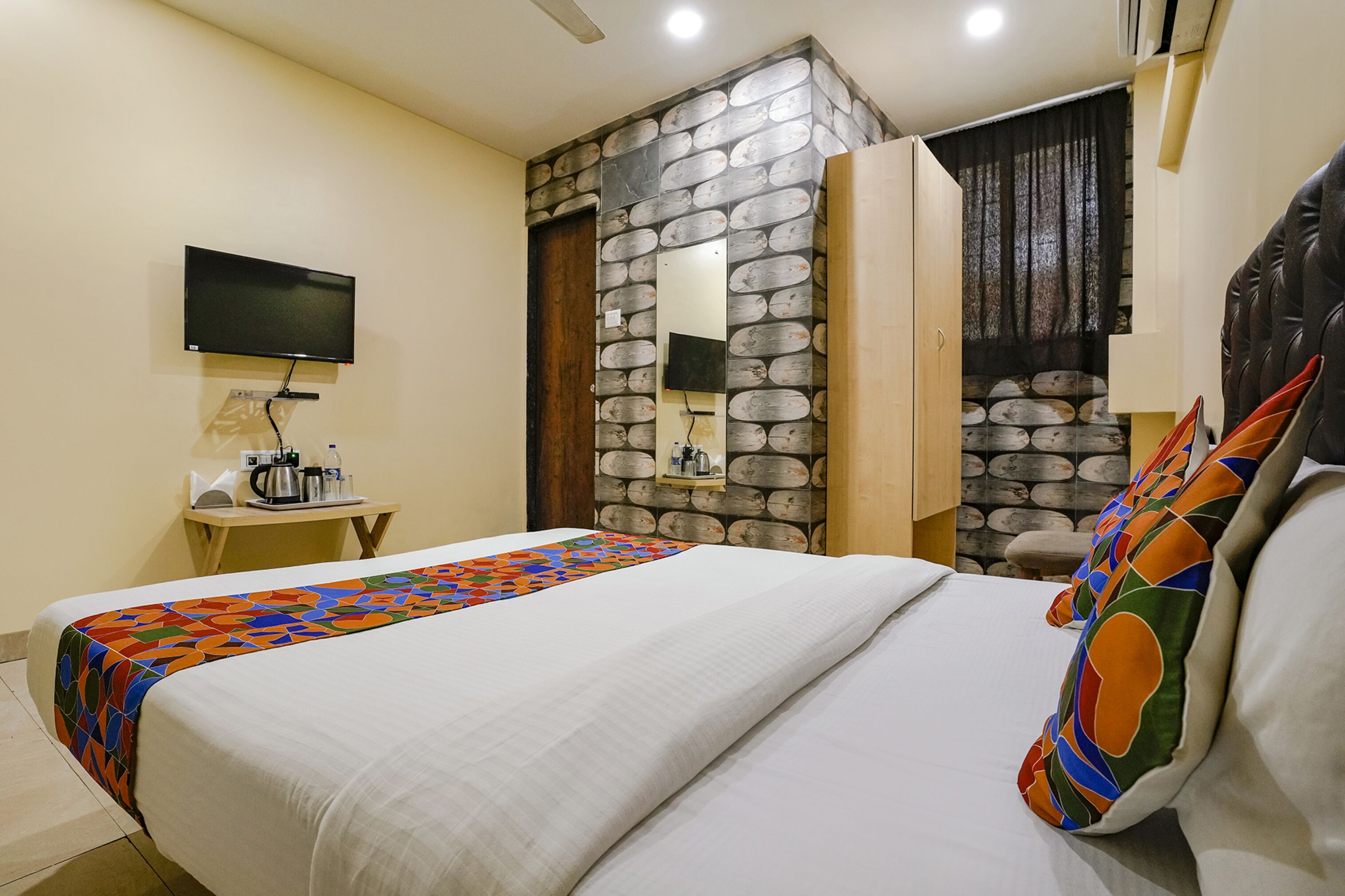 A hotel room with a bed, a TV mounted on the wall, a small table with a kettle, and a stone wall.