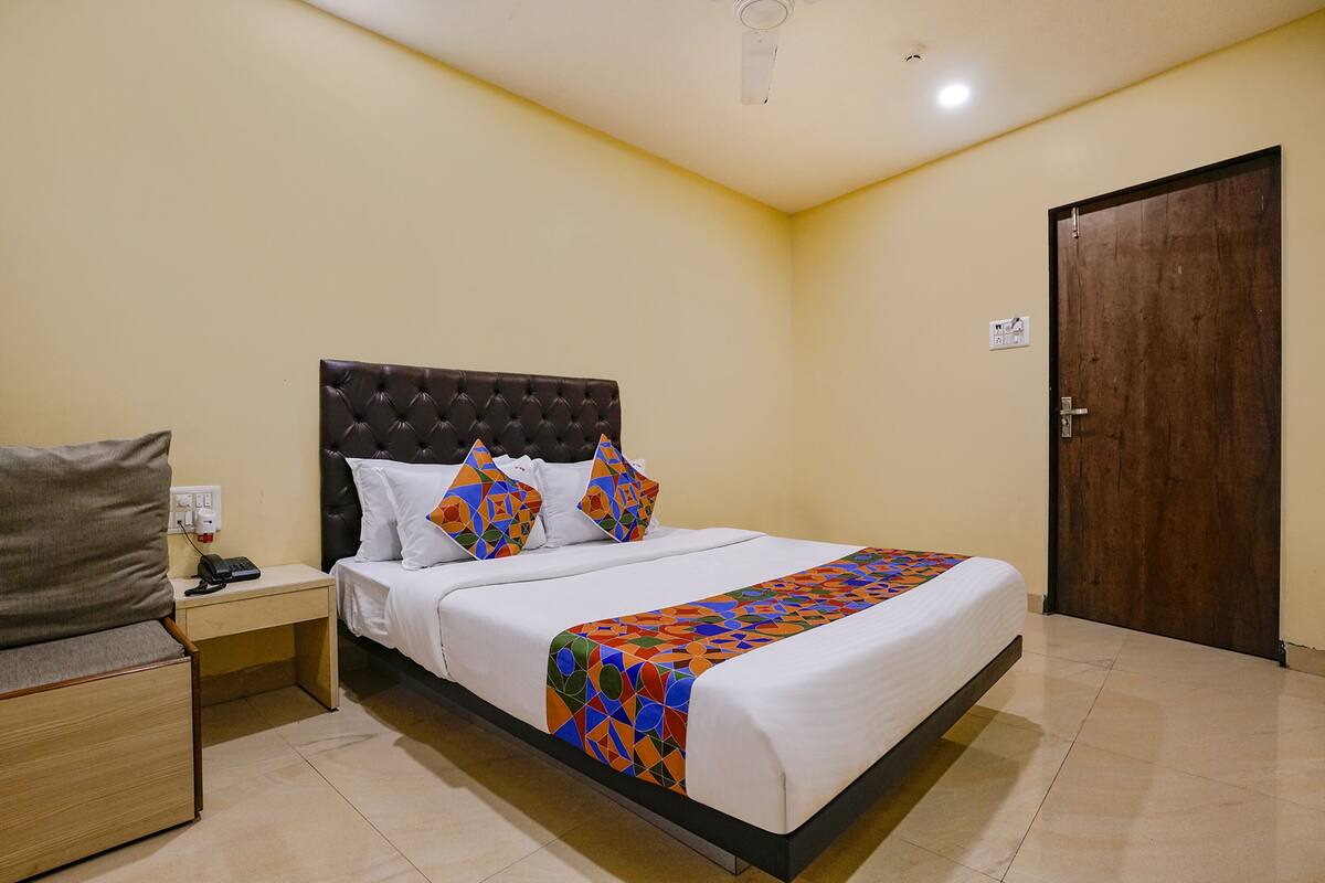 Una cama bien hecha con una colcha estampada, una mesita de noche con un teléfono y una puerta de madera en una habitación con paredes color crema.