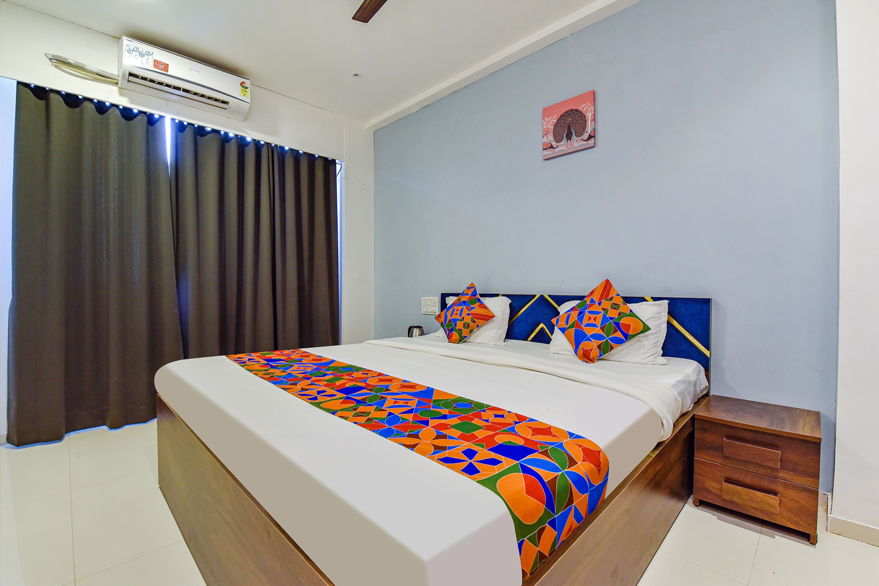 A hotel room with a bed, a nightstand, a wall-mounted air conditioner, and a window with curtains.