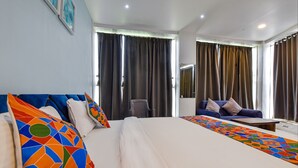 Habitación de hotel con una cama grande, un sofá, una mesita y una silla. Hay cortinas en las ventanas y un cuadro en la pared.