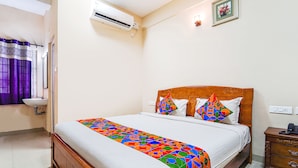 A neatly made bed with a colorful bedspread, a wooden headboard, and two pillows. There is a small wooden nightstand with a phone and a wall-mounted air conditioner above the bed. - Fabexpress Uv Residency (Coimbatore)