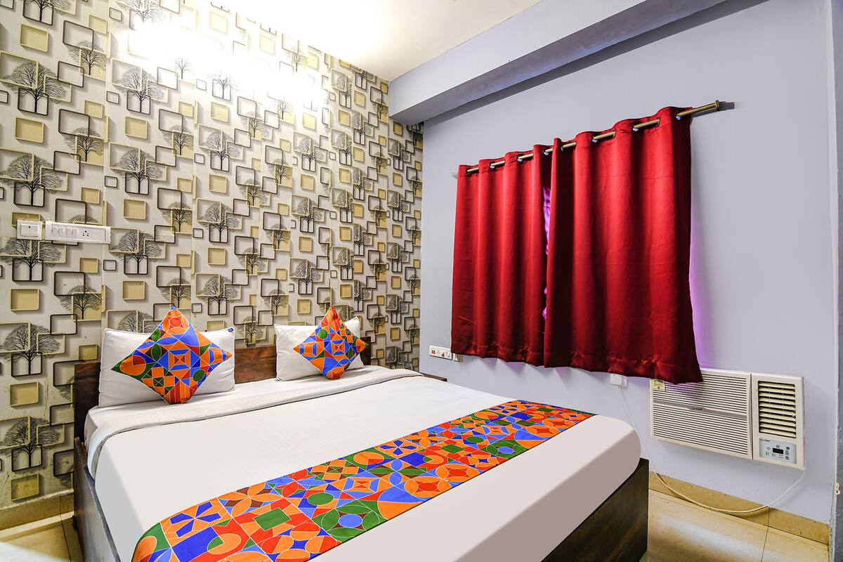 Een hotelkamer met behang met een patroon, een bed met kleurrijke kussens, een rood gordijn en een luchtverkoeler aan de muur.