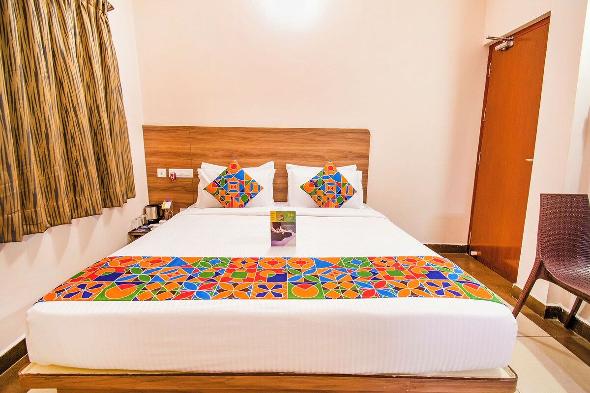 Habitación de hotel con cabecera de madera, cama con ropa blanca y almohadas de diseño geométrico colorido, una mesita con un libro y una puerta de madera.