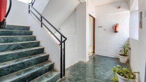 Una escalera con peldaños de mármol verde, barandilla negra y pared blanca con un extintor y una planta en maceta.