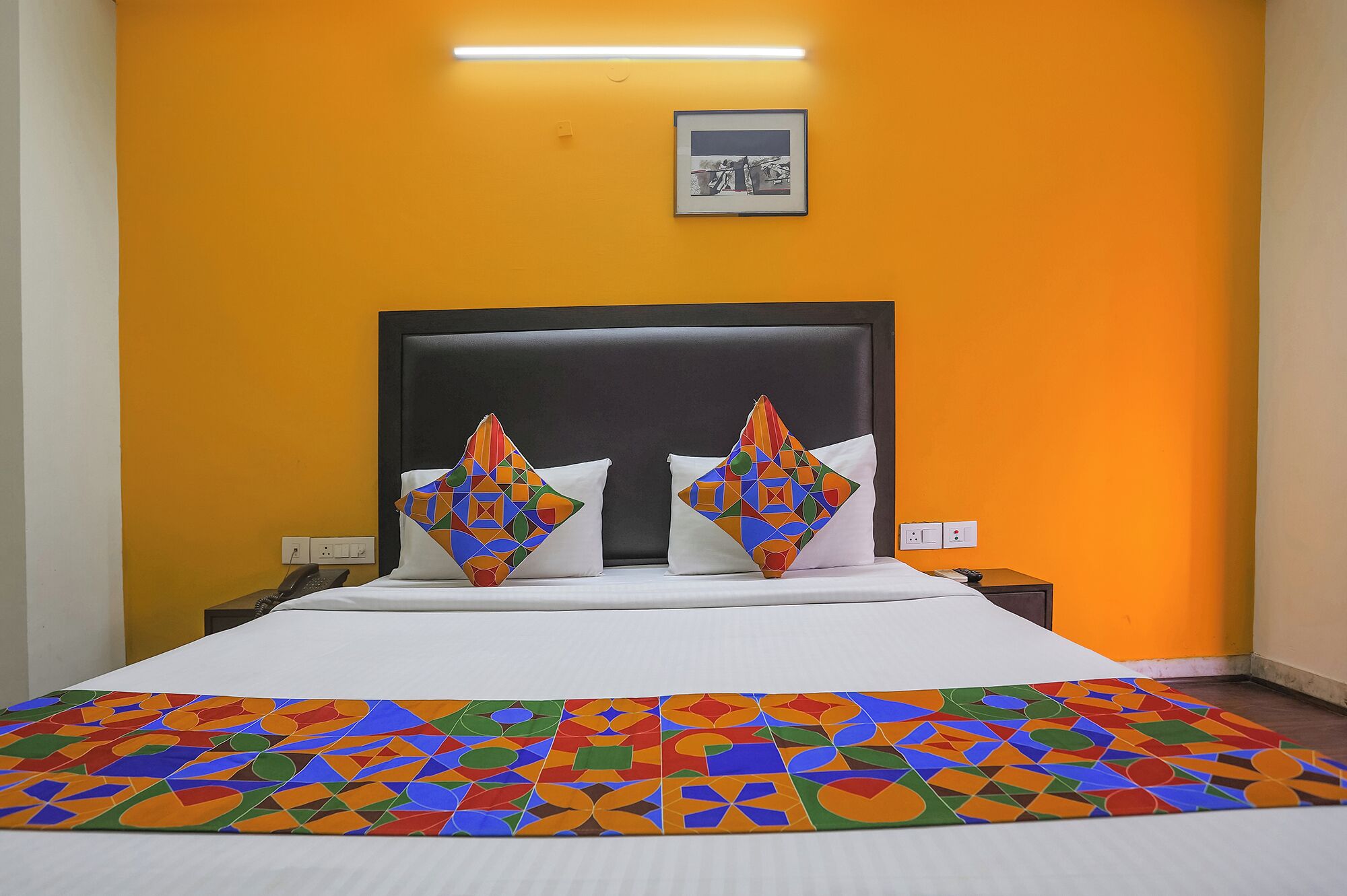 Une chambre d’hôtel avec un lit, deux oreillers à motifs géométriques, un tableau encadré au mur et un tapis coloré.
