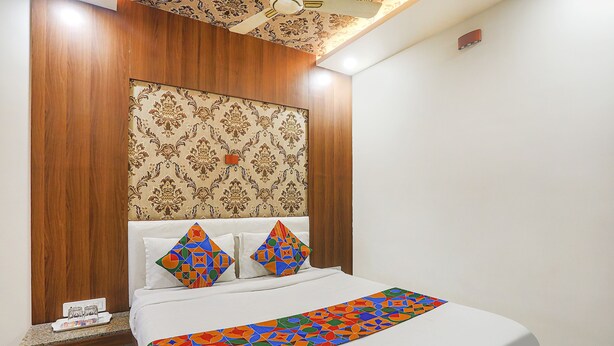 A neatly made bed with a colorful geometric-patterned bedspread, a wooden headboard with a decorative panel, and a small bedside table with a phone and a glass. - Fabexpress Jai Manglam Service Apartment (Nagpur)