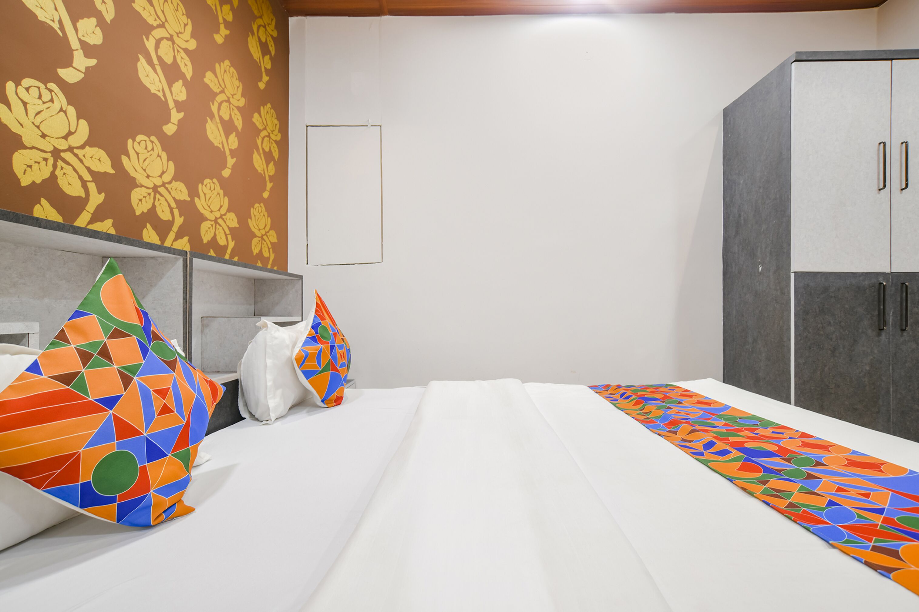A hotel room with a bed, colorful pillows, a patterned wall, and a wardrobe.