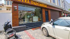 Um hotel chamado "HOTEL TAJ CASTLE" com fachada moderna, entrada visível e um carro e uma scooter estacionados na frente.