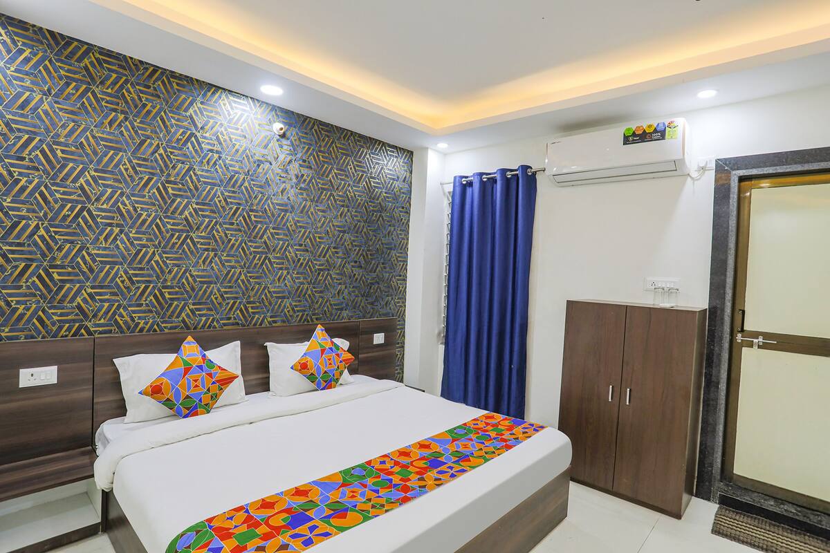 A hotel room with a patterned wall, a bed with colorful pillows, a blue curtain, a wardrobe, and an air conditioning unit.