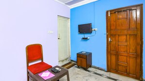 A room with a wooden door, a television mounted on the wall, a small wooden cabinet, a red chair, and a table with a remote control and a pink folder. - Fabexpress Karunamoyee Regency (Barasat)