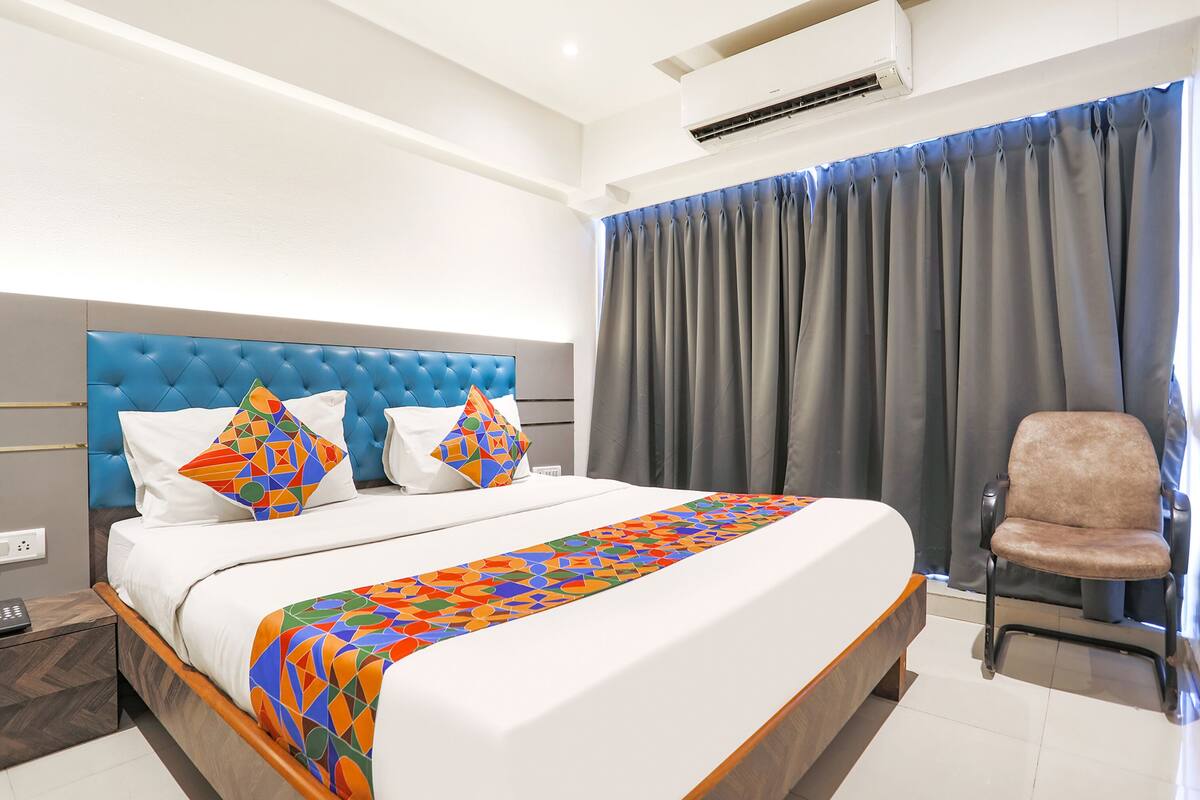 A neatly made bed with a geometric-patterned throw, a tufted headboard, and a chair beside a window with grey curtains.
