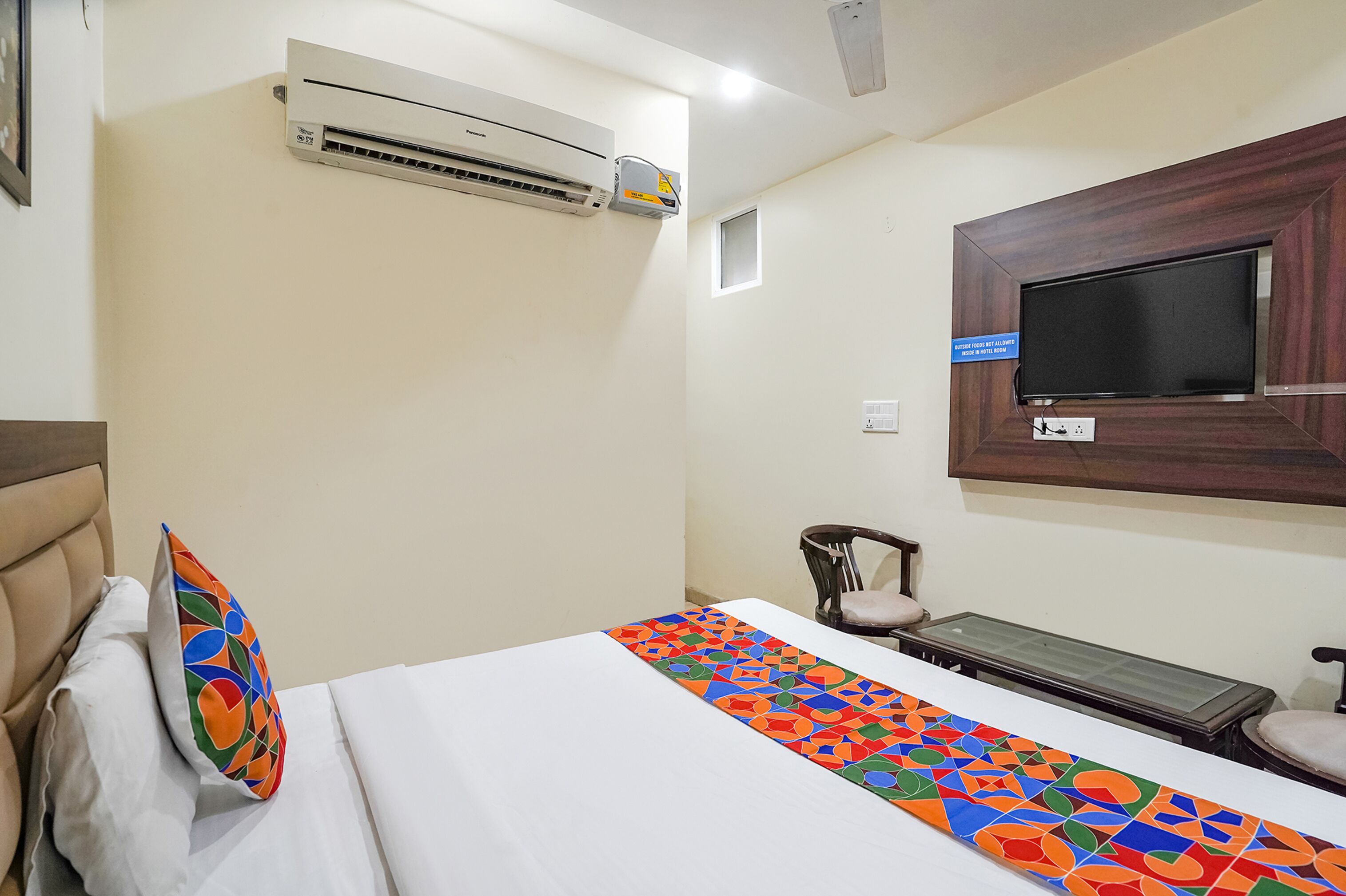 A hotel room with a bed, a TV mounted on the wall, an air conditioning unit, and a small table.