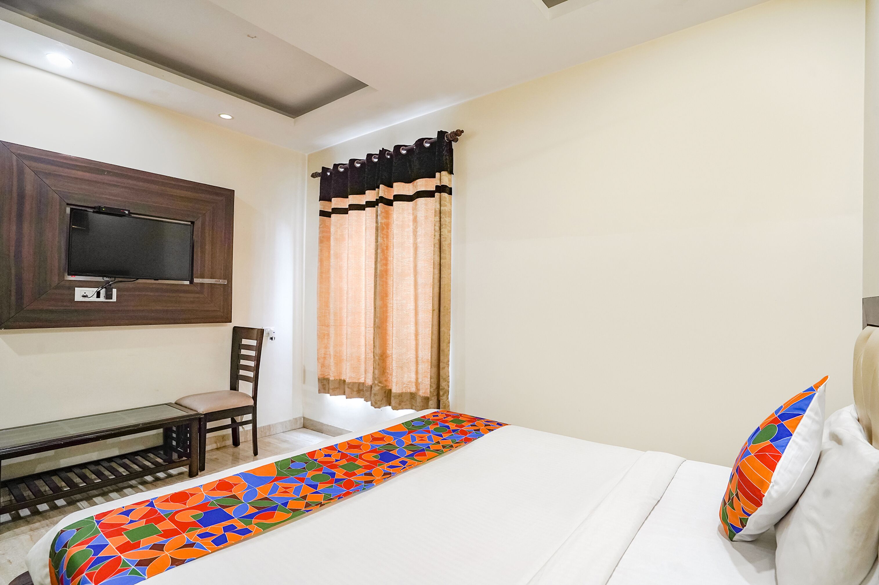 A hotel room with a bed, a TV mounted on the wall, a bench, and a window with curtains.