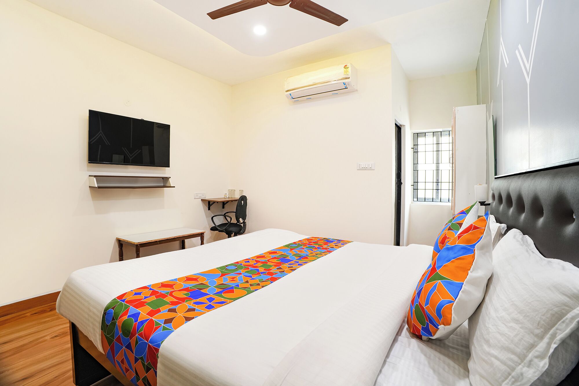 A hotel room with a bed, a TV mounted on the wall, an air conditioning unit, a desk, and a chair.
