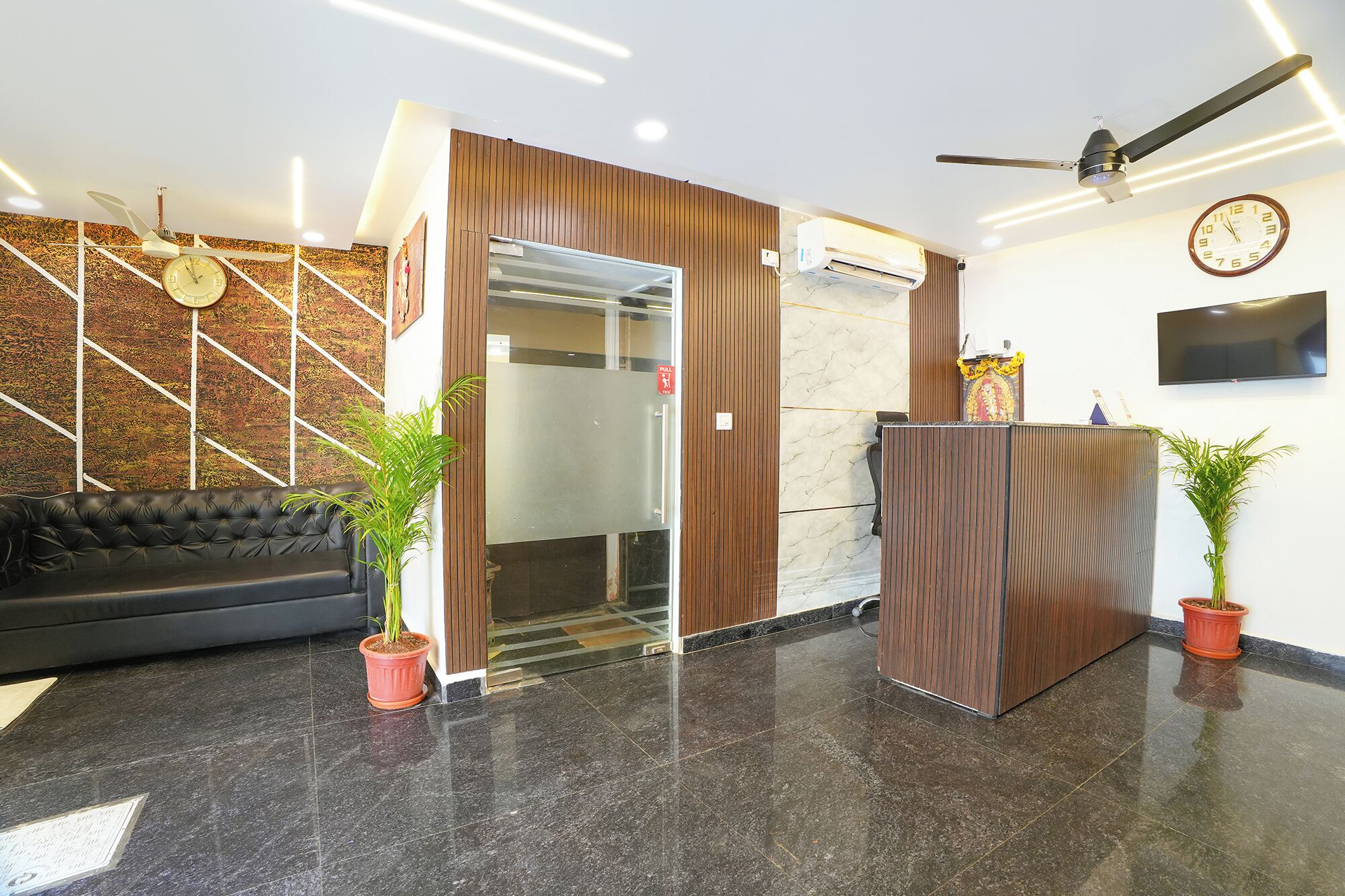 A reception area with a black leather sofa, two potted plants, a wooden reception desk, a wall clock, and a ceiling fan.