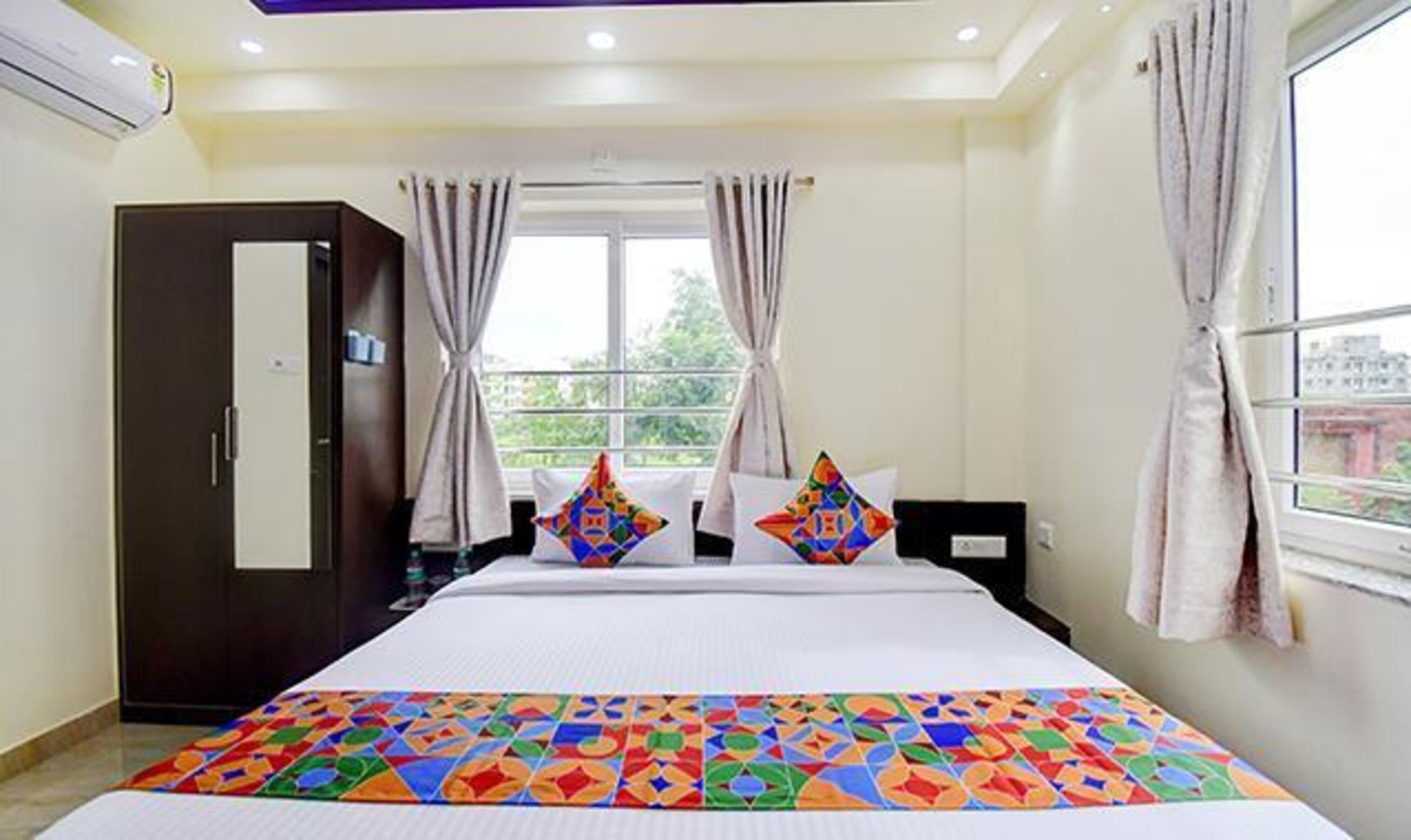 A hotel room with a large bed, a wardrobe, a window with curtains, and a view of greenery outside.