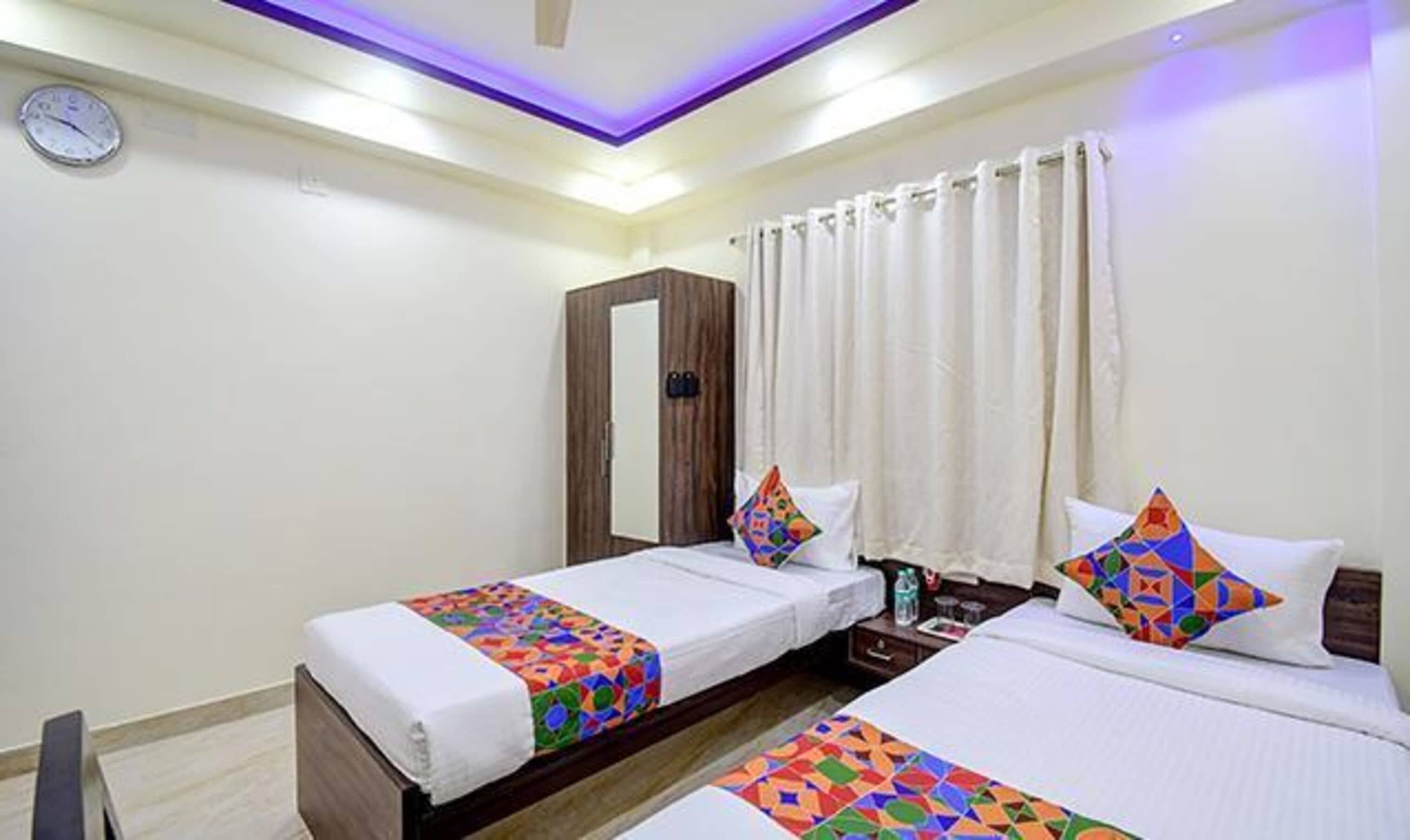 A hotel room with two beds, a mirror, a clock, and a window with curtains.