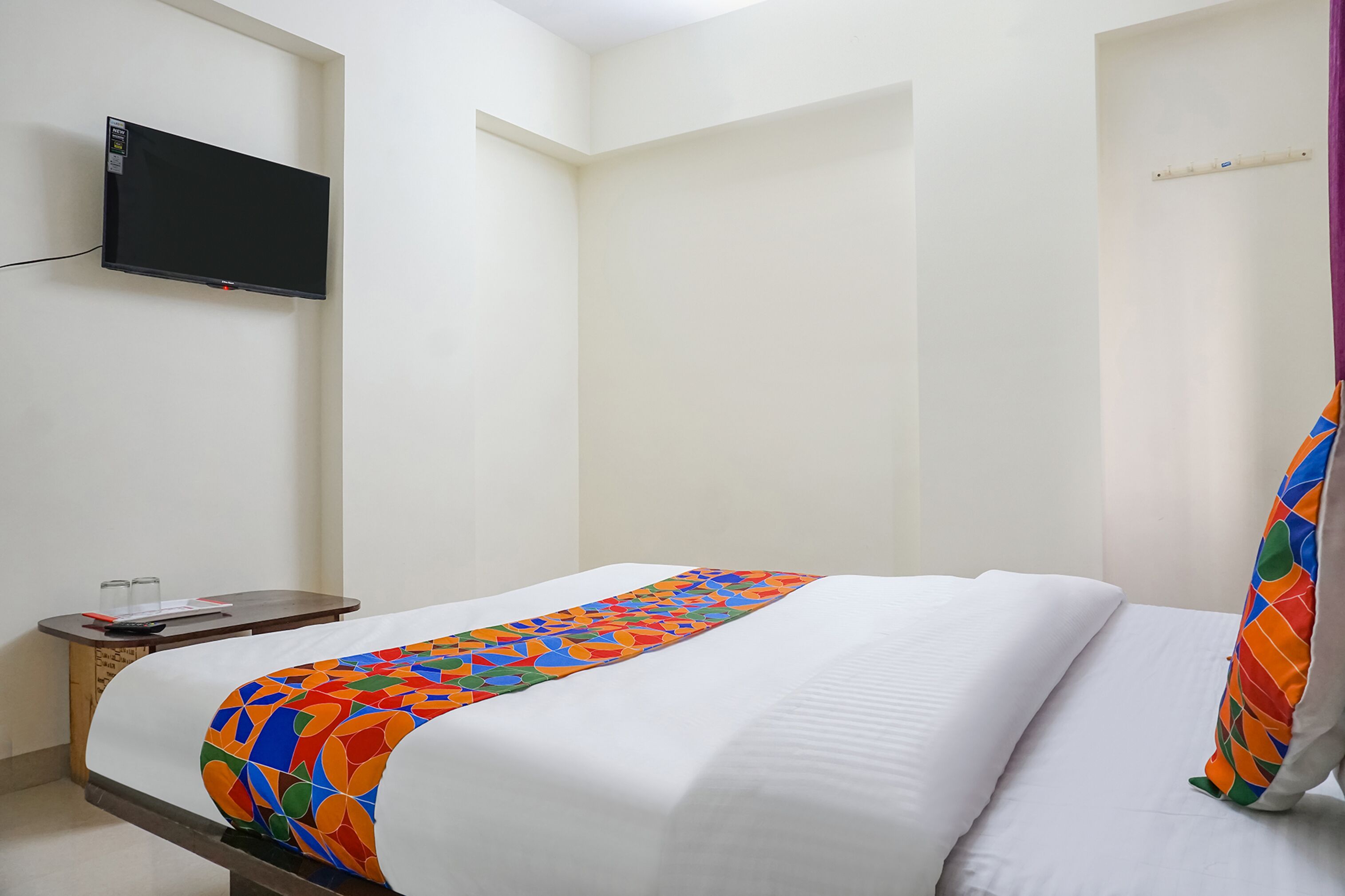 A hotel room with a bed, a TV mounted on the wall, a small table with glasses and a notebook, and a colorful bedspread.