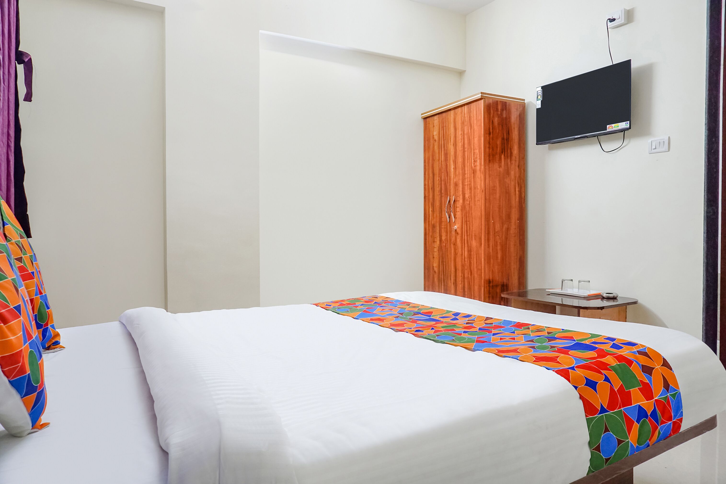 A hotel room with a bed, a wooden wardrobe, a television mounted on the wall, and a small table with a lamp.