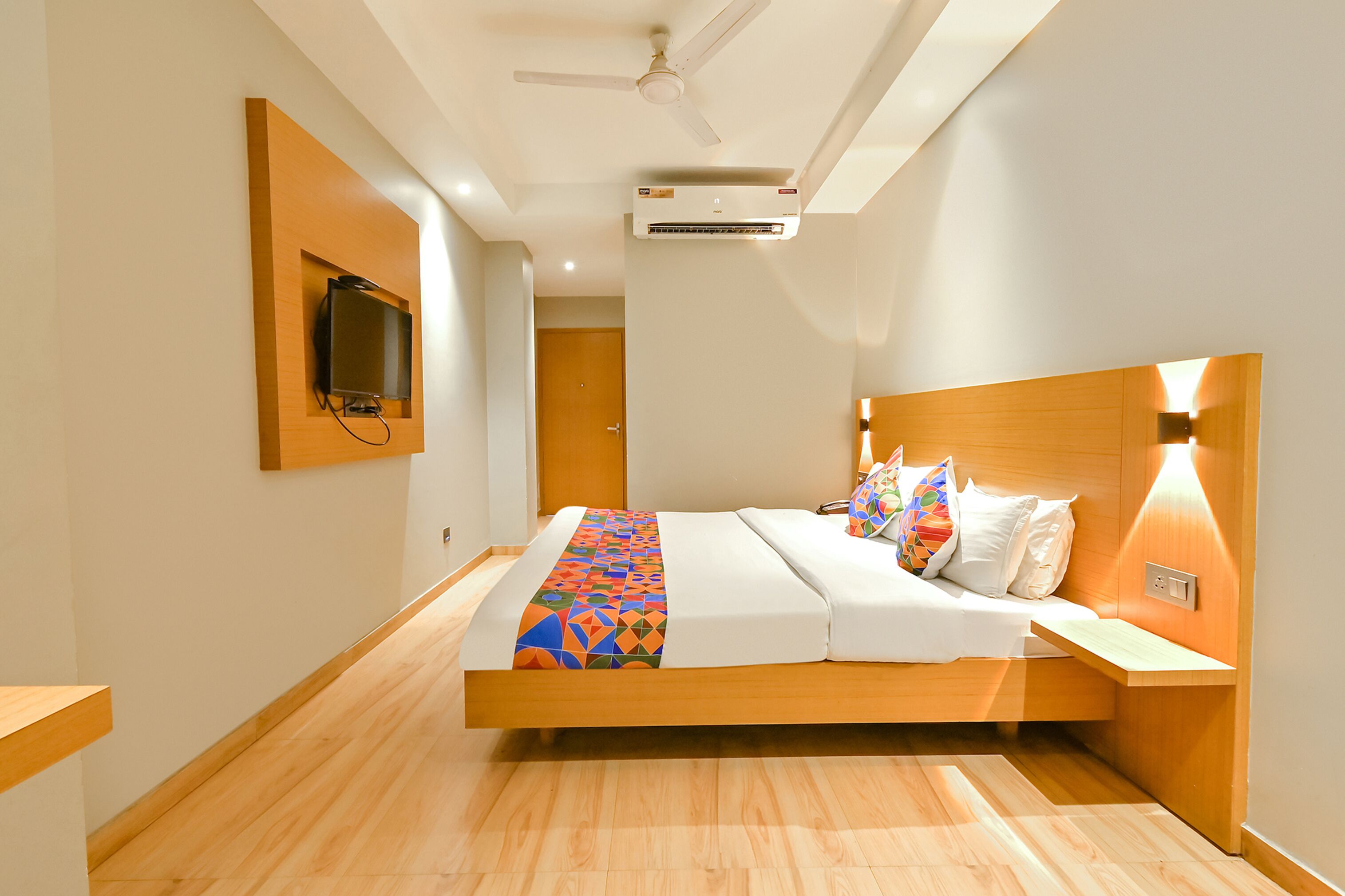 Una habitación de hotel moderna con una cama de madera, televisor montado, equipo de aire acondicionado y ventilador de techo.