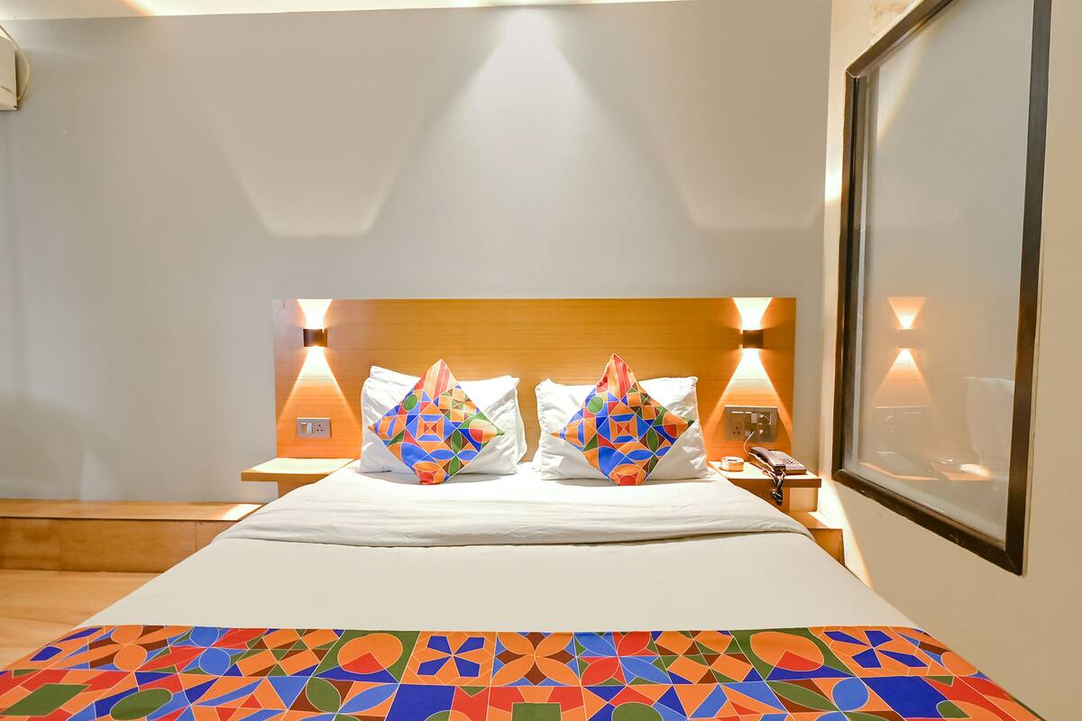 A hotel room with a wooden headboard, two bedside lamps, a bed with white bedding, and a colorful geometric-patterned throw.