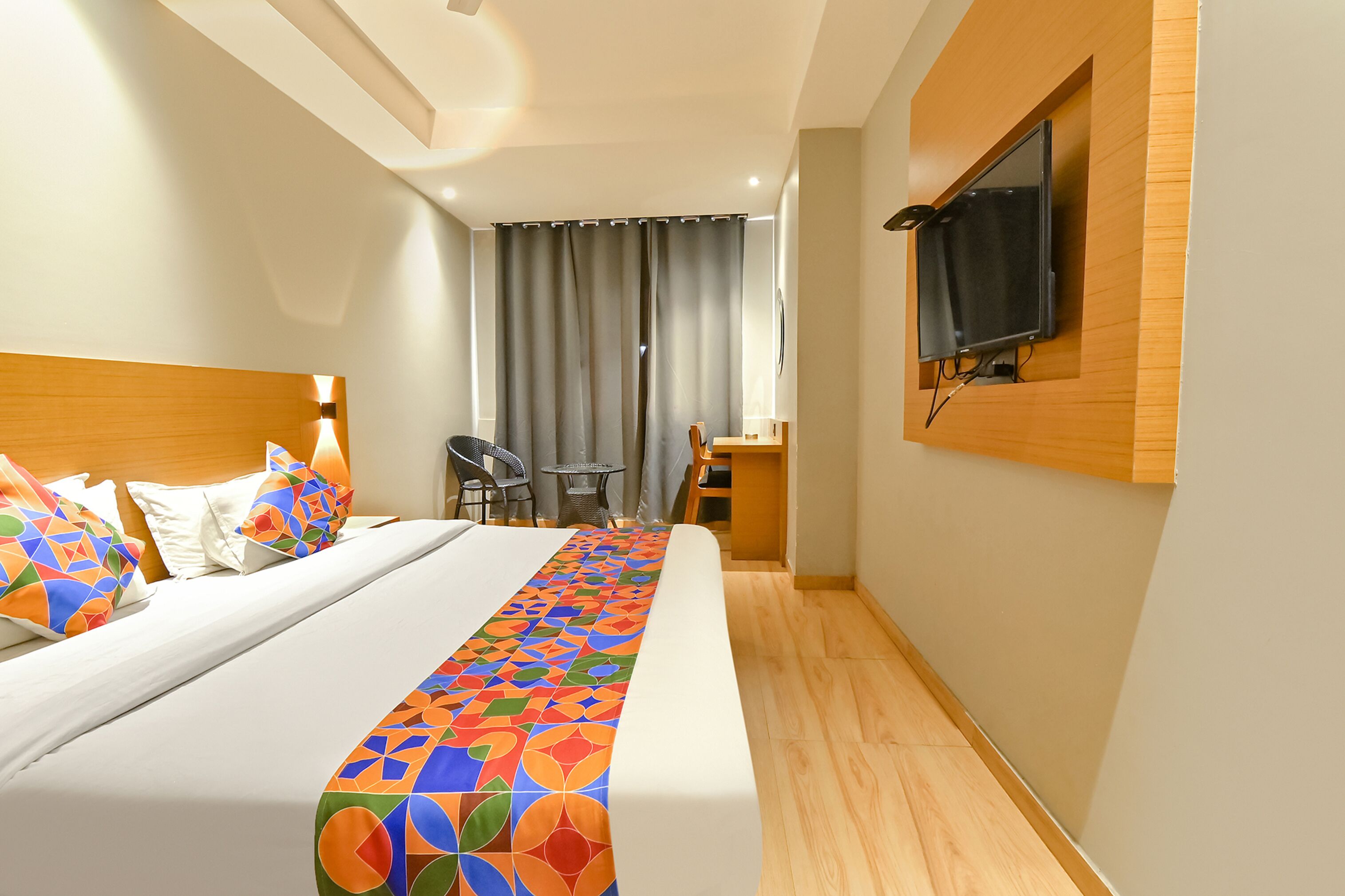 A hotel room with a bed, a TV mounted on the wall, a desk with a chair, and a window with curtains.