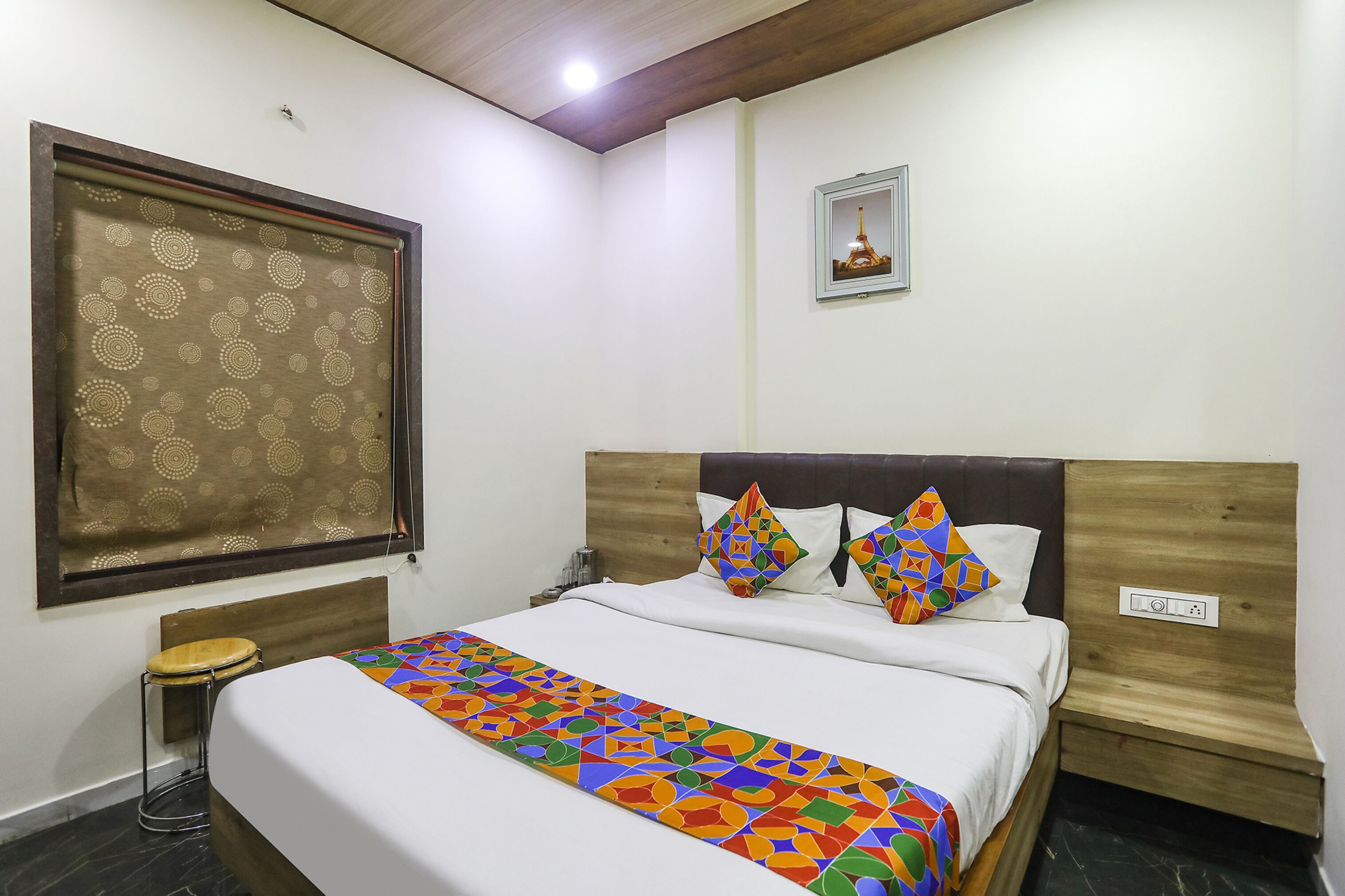 A neatly arranged hotel room with a bed, a small stool, a nightstand, and a wall-mounted picture.
