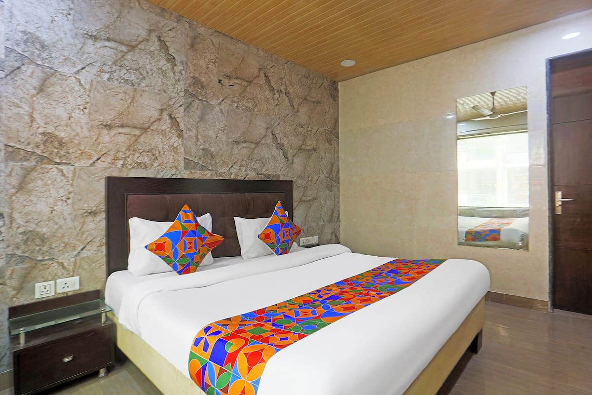 A hotel room with a stone wall, a wooden ceiling, a bed with white bedding, colorful pillows, a glass bedside table, and a window with a view.