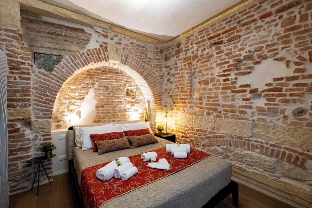 Ein Zimmer mit Backsteinwand, einem Bett mit roter und beiger Bettdecke sowie einem kleinen Tisch mit Lampe.
