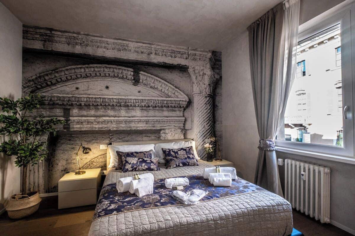 Ein Schlafzimmer mit einem Stein-Kopfteil, einem Bett mit floralem Bettzeug, einem Nachttisch mit Lampe und Blick auf die Stadt durch ein Fenster.