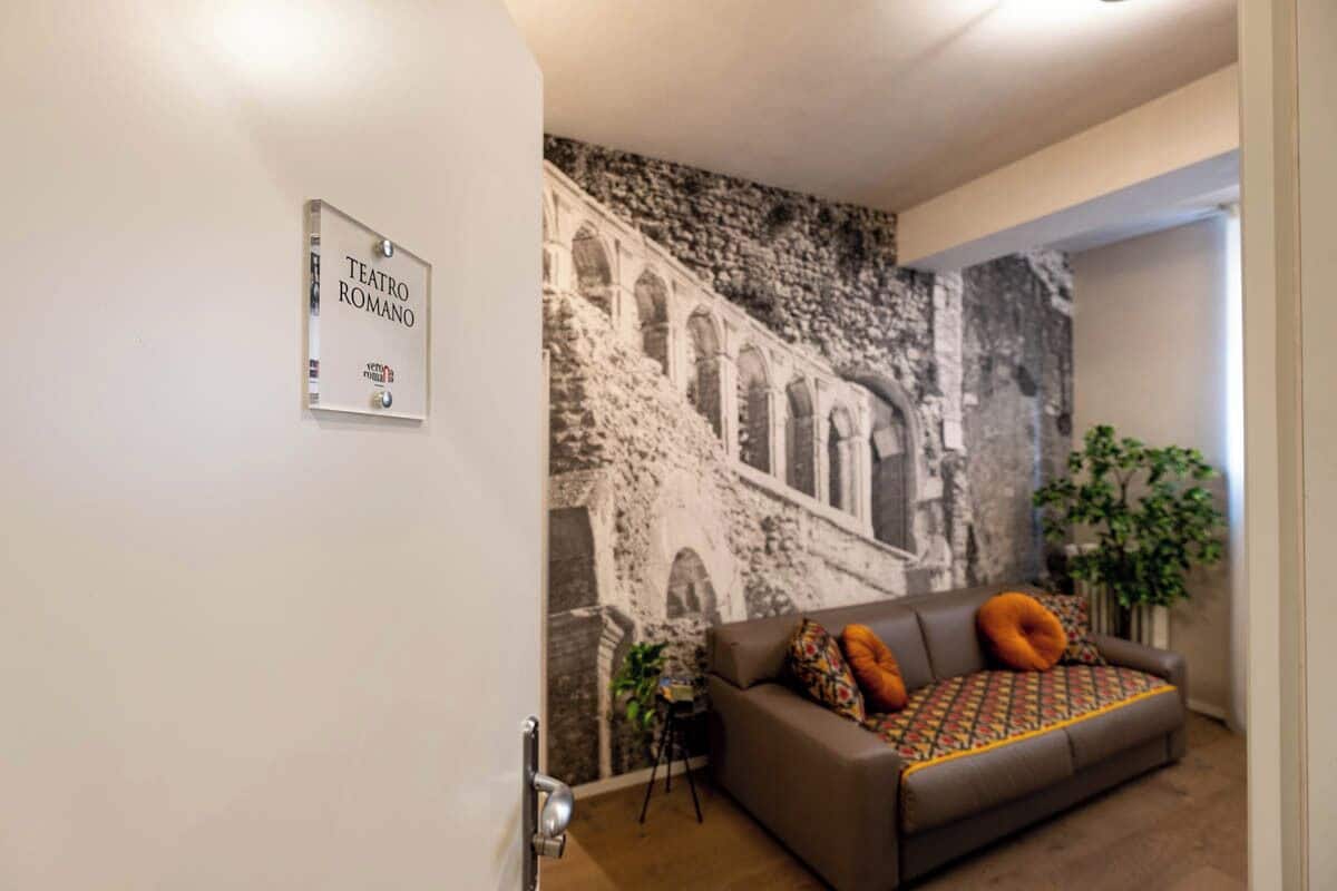 Ein Wohnzimmer mit Wandgemälde einer antiken Struktur, einem braunen Sofa mit farbenfrohen Kissen und einem Schild mit der Aufschrift 'TEATRO ROMANO'.