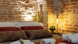 Ein gemütliches Zimmer mit Backsteinwand, einem Bett mit roten und beigen Kissen, einer kleinen Pflanze sowie ordentlich gefalteten Handtüchern und Flaschen auf dem Boden.