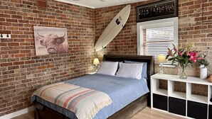 Une chambre avec un mur en briques, un lit recouvert d’une couverture bleue, un ventilateur de plafond et une planche de surf accrochée au mur.