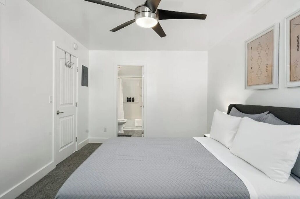 A bedroom with a bed, a ceiling fan, a white door, and a bathroom visible through an open door.